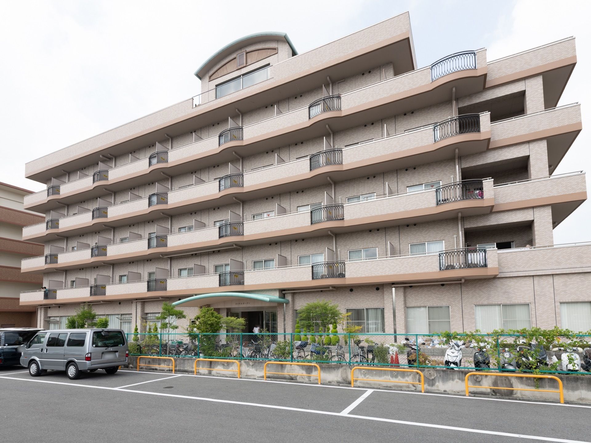 薄いピンク色の外壁に、各居室にはベランダが用意されている。１階には駐輪場があり、自転車や原付自動車が停まっている。