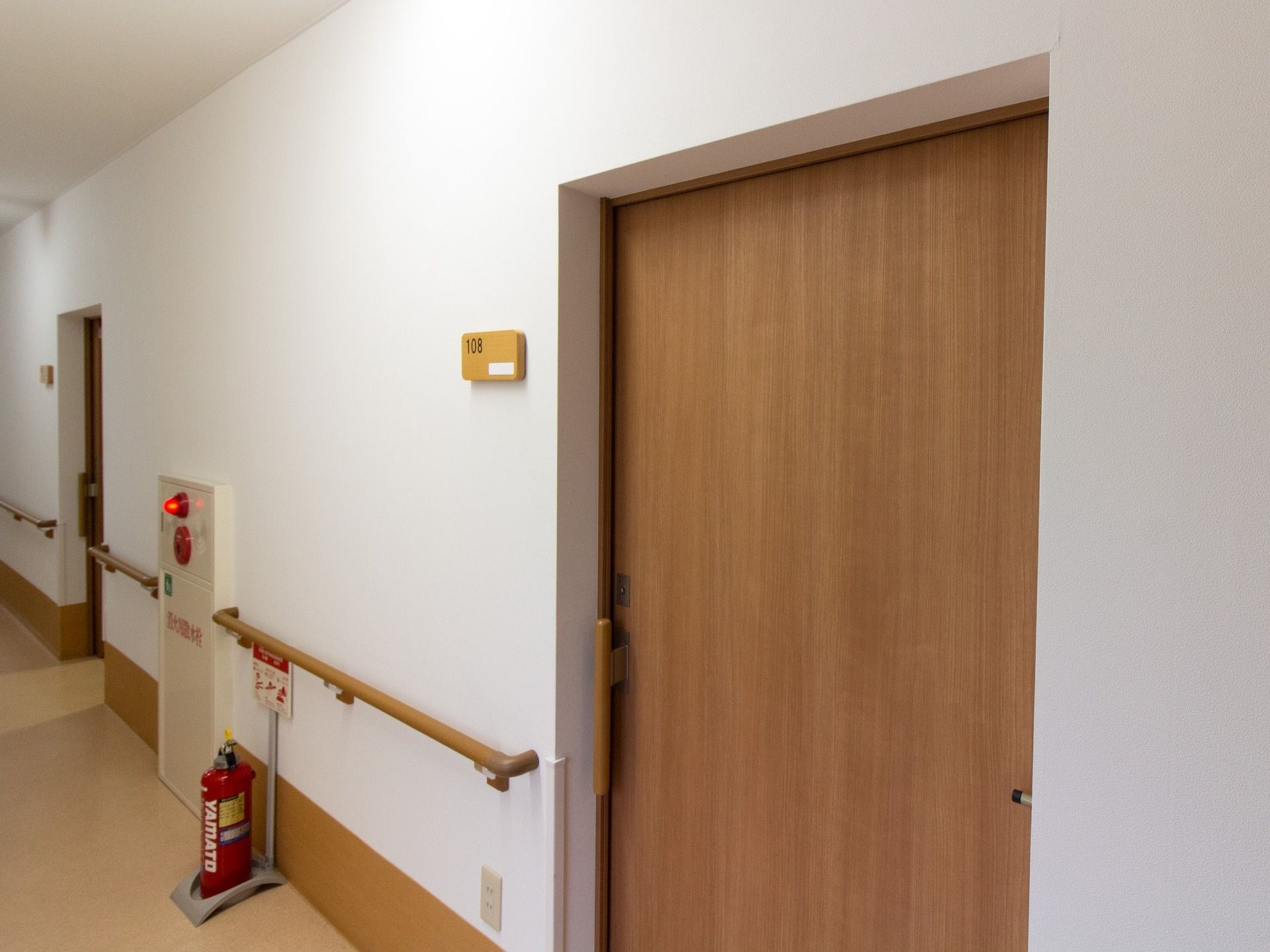 廊下に居室への木製ドアがある。引き戸になっており、扉の横の壁にネームプレートが設置されている。奥に消火器が置いてある。