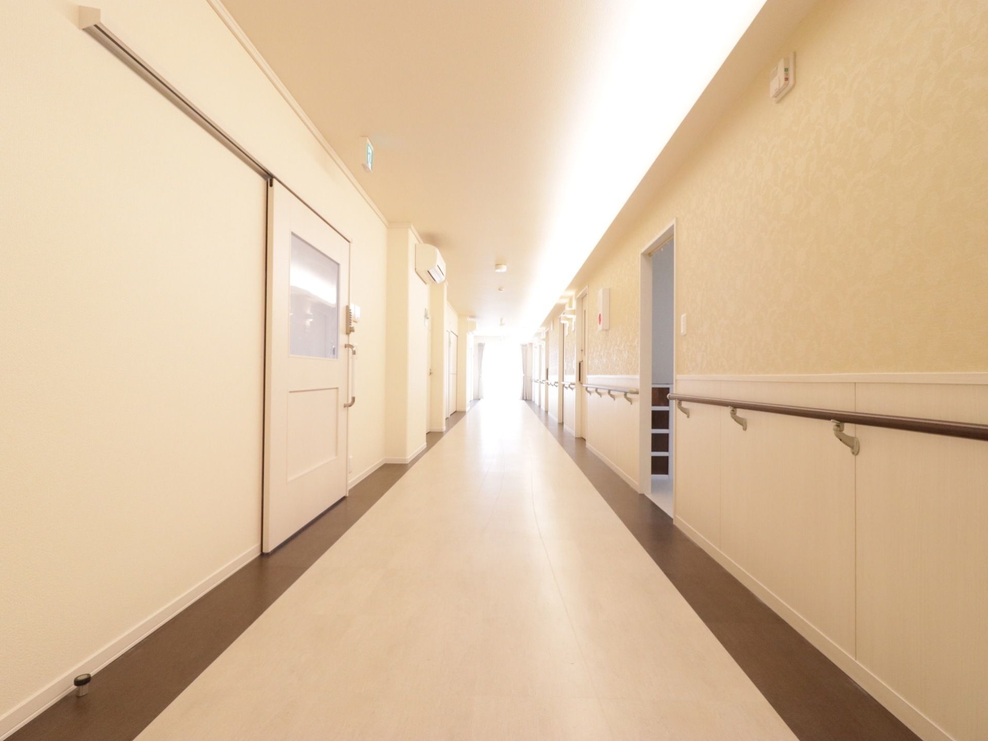 突き当りに窓のあるまっすぐな通路である。光が入っていて、床に反射している。壁にエアコンが設置されている。