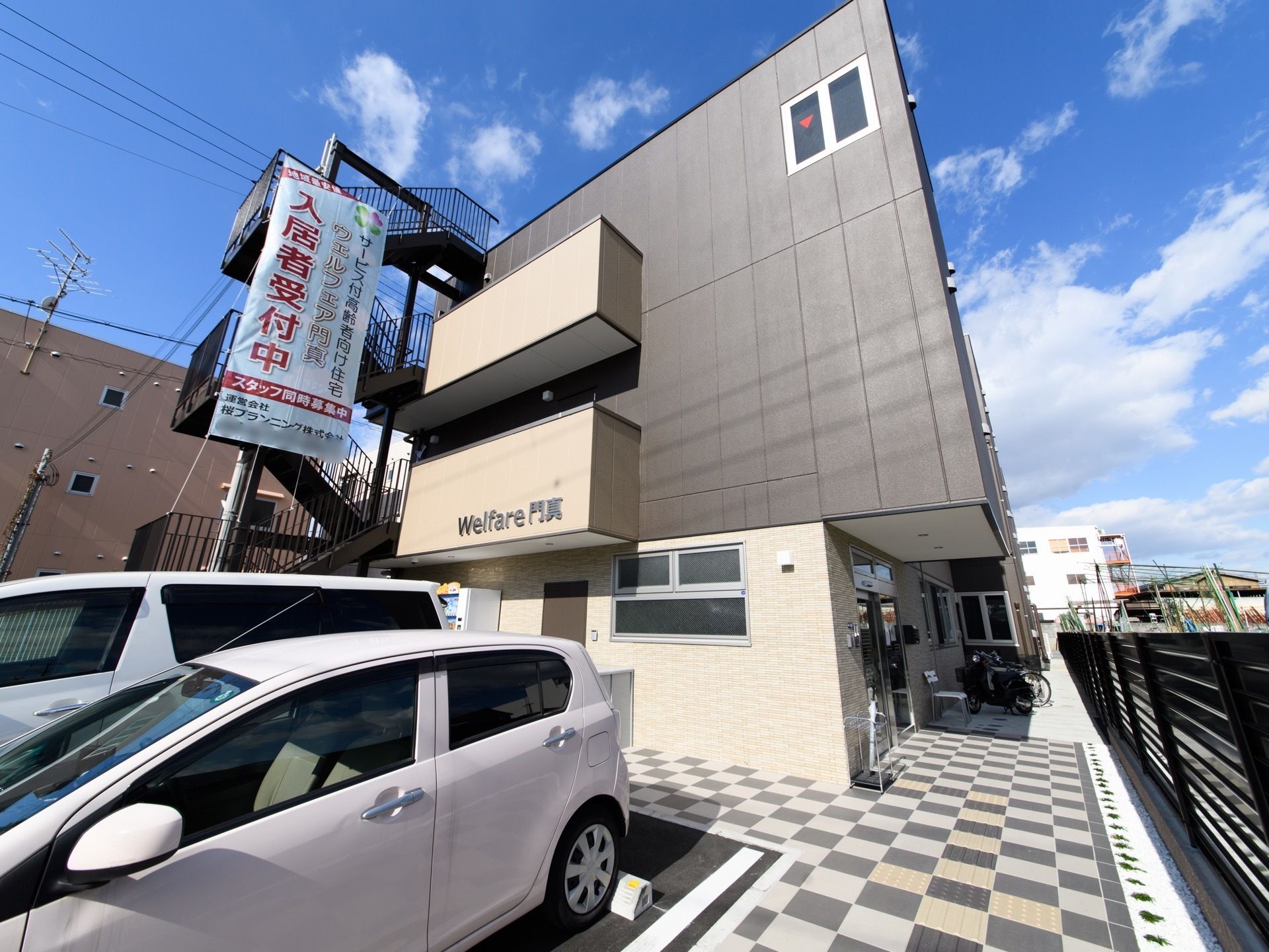 3階建てのモダンスタイルの建物である。市松模様に敷かれているタイルがおしゃれである。建物周辺はバリアフリ―である。