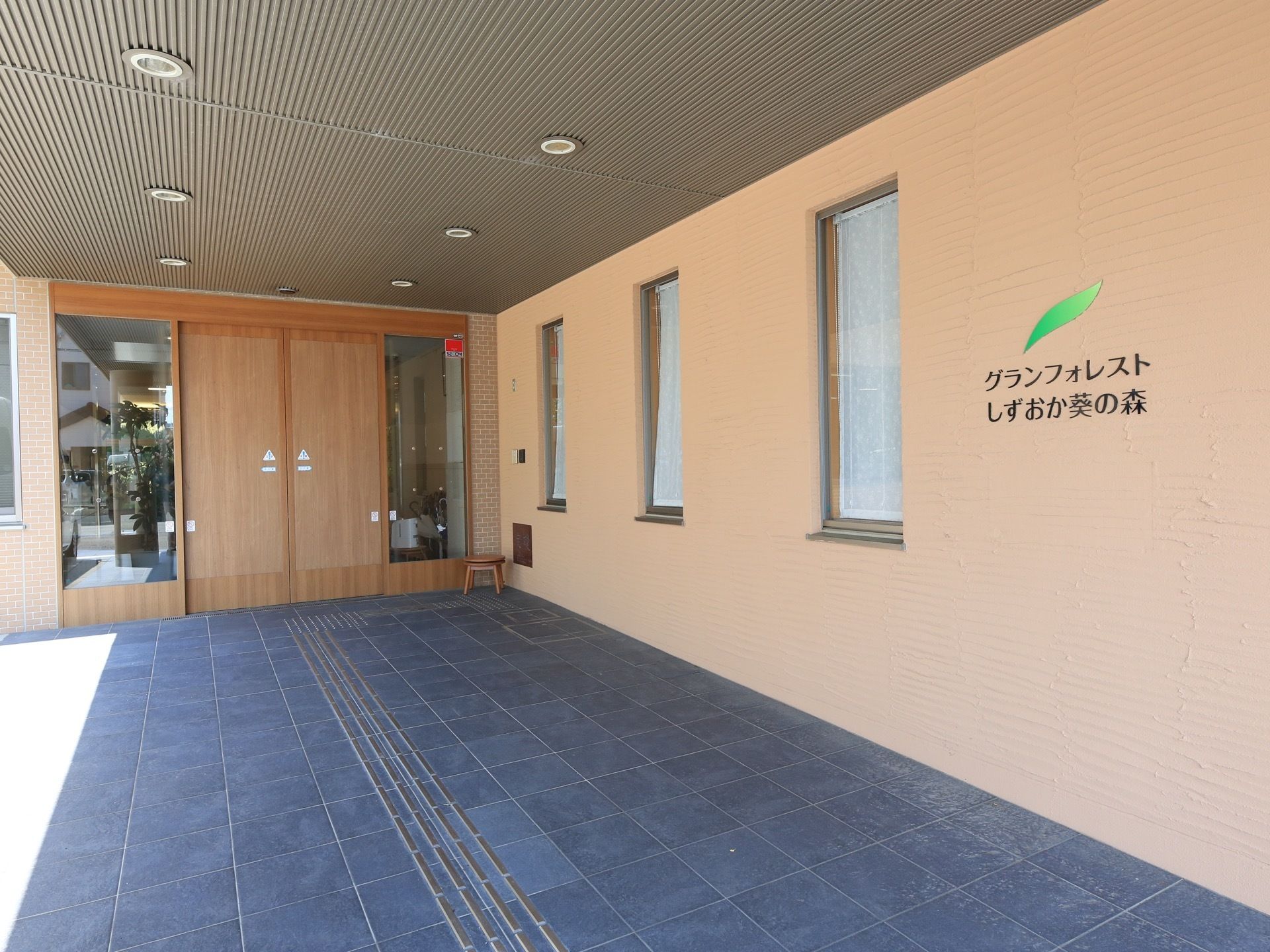 玄関には木目調の自動ドアが設置されている。ベージュ色の壁に縦長の窓が３つあり、その横に施設名が表示されている。
