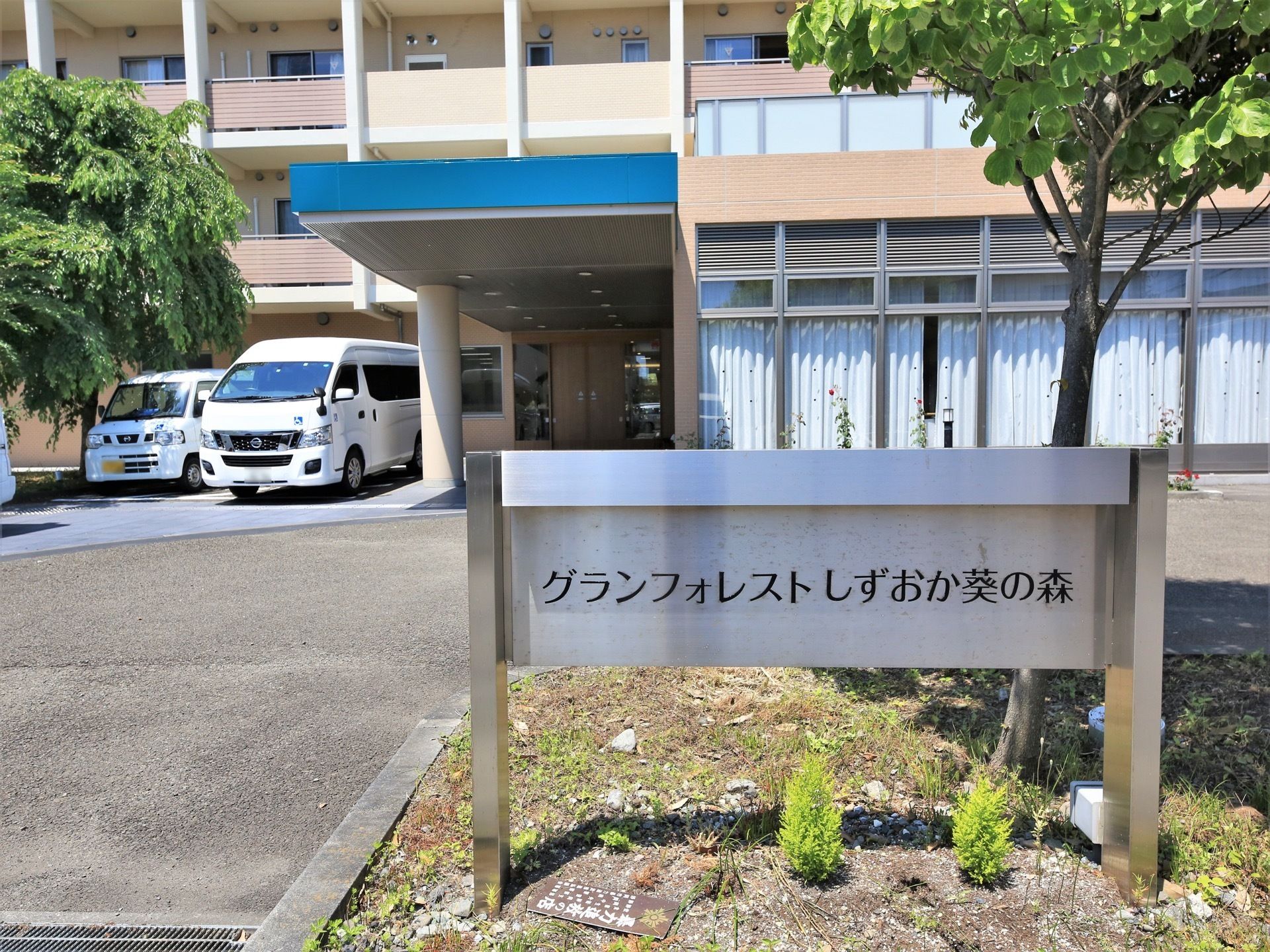 建物の前方に植え込みがあり、樹木が植えられている。植え込みの中に銀色の施設名が書かれた看板が立っている。