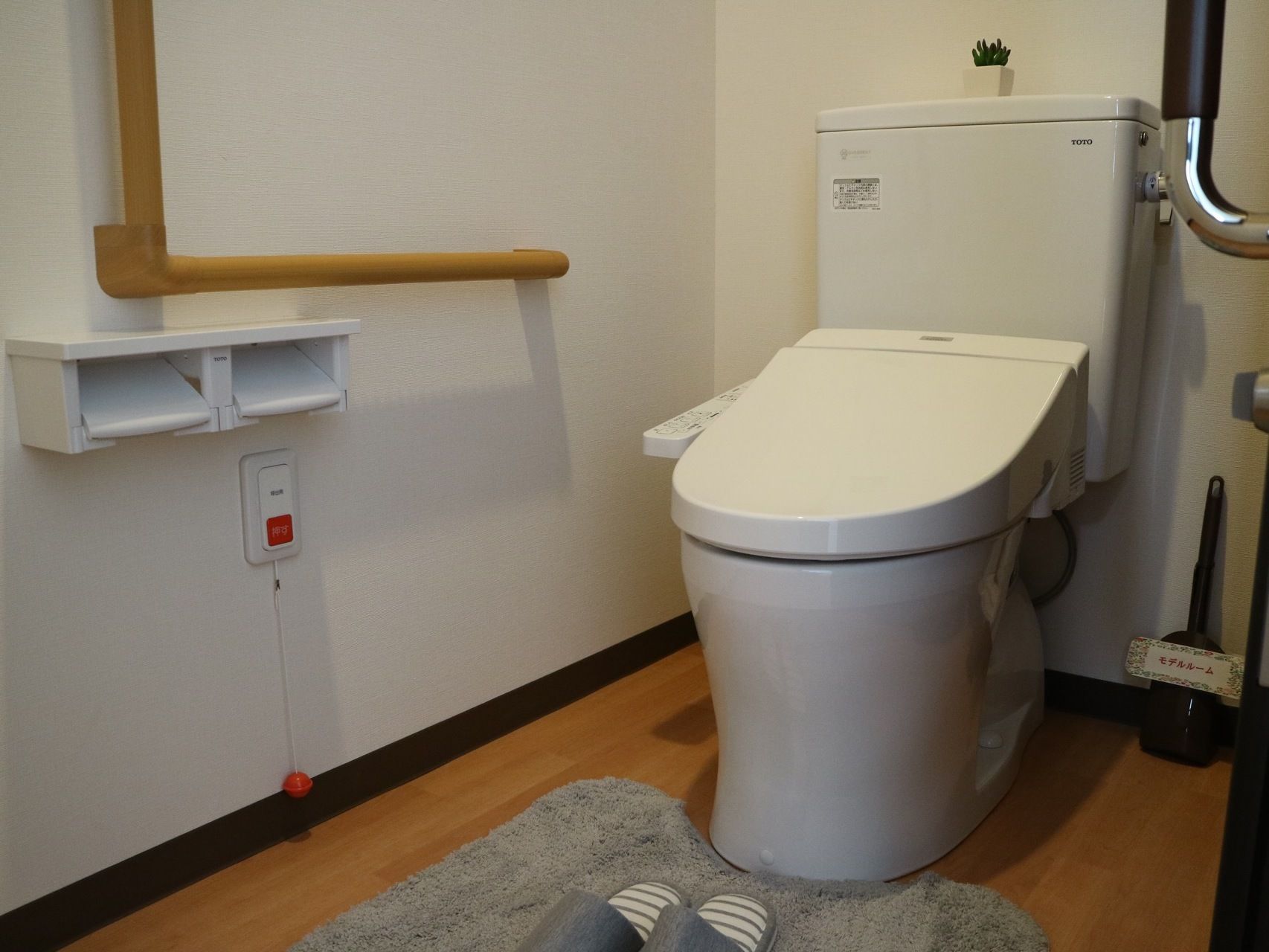 居室には、温水洗浄便座付きのトイレが完備されている。L型手すりと呼び出しボタンが壁に設置されている。