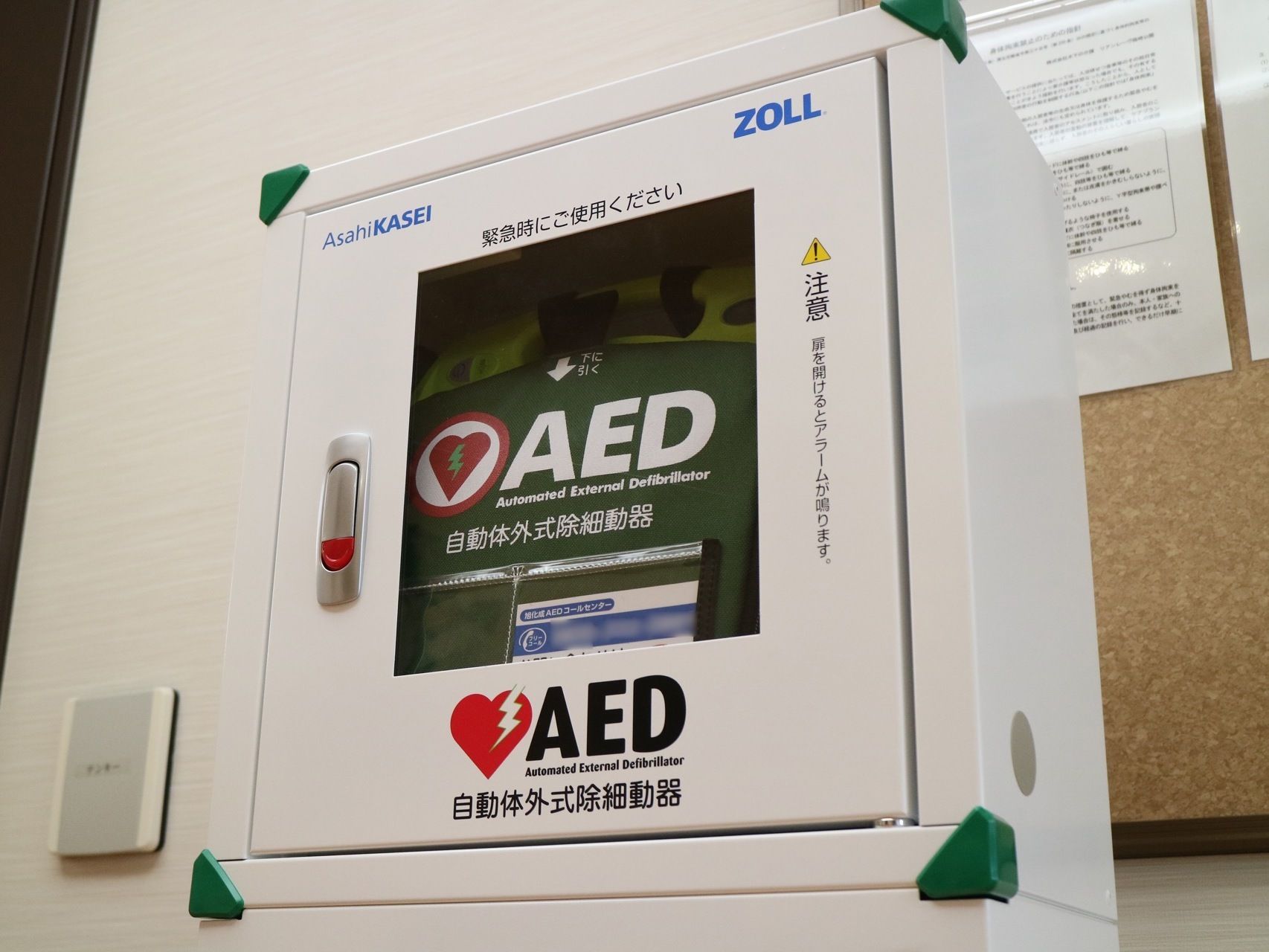 エントランスホールの壁際には、AEDが設置がされている。コルクボードが後ろの壁に取りつけられている。