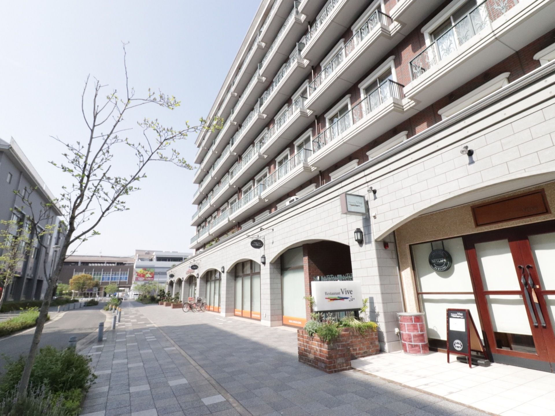 建物は洋風なデザインで１階部分は店舗となっている。歩道と車道が区分けされており、樹木が植えられている。