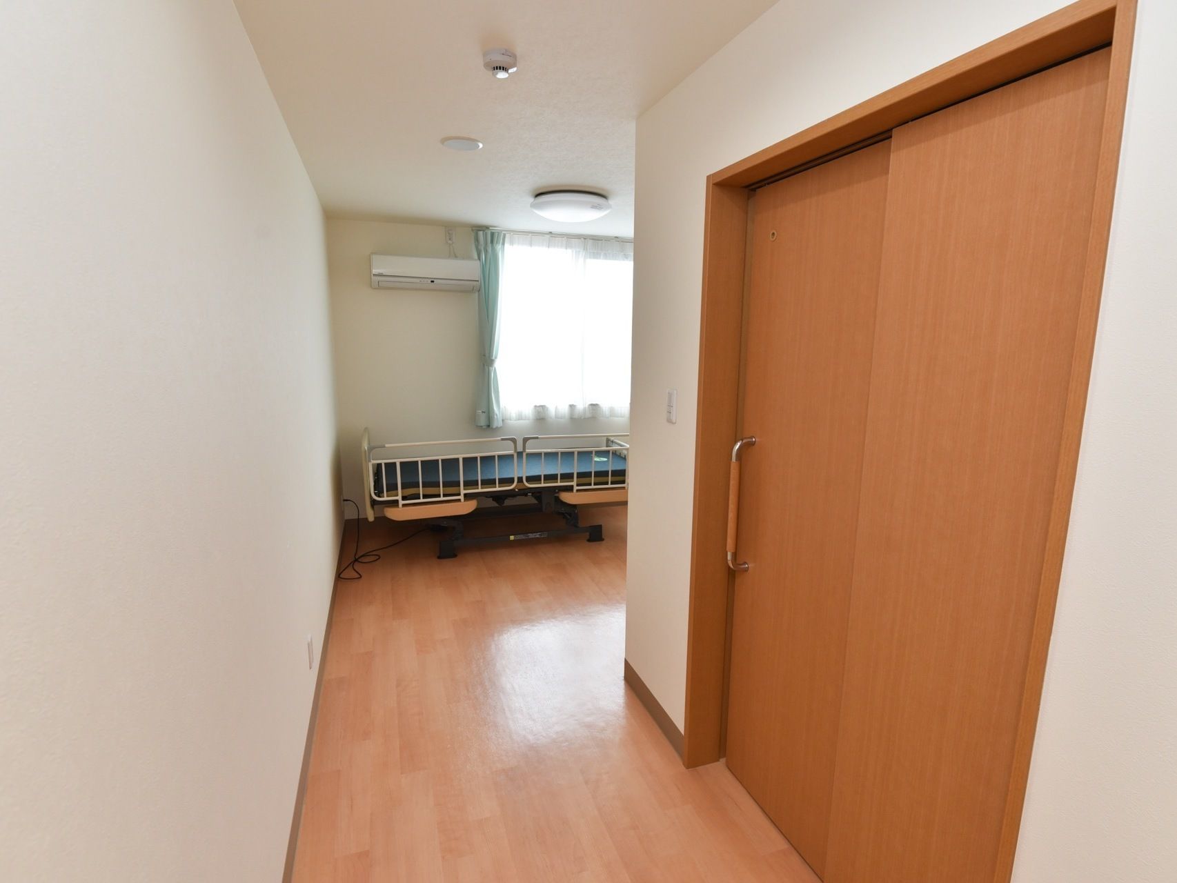 居室はフローリングになっている。介護用ベッドやエアコン、煙探知器が設置されている。大きな横長の窓がある。