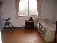 洋室タイプの居室である。介護ベッドと収納家具が配置されており、歩行車が置かれている。カーテンが掛けられた窓がある。
