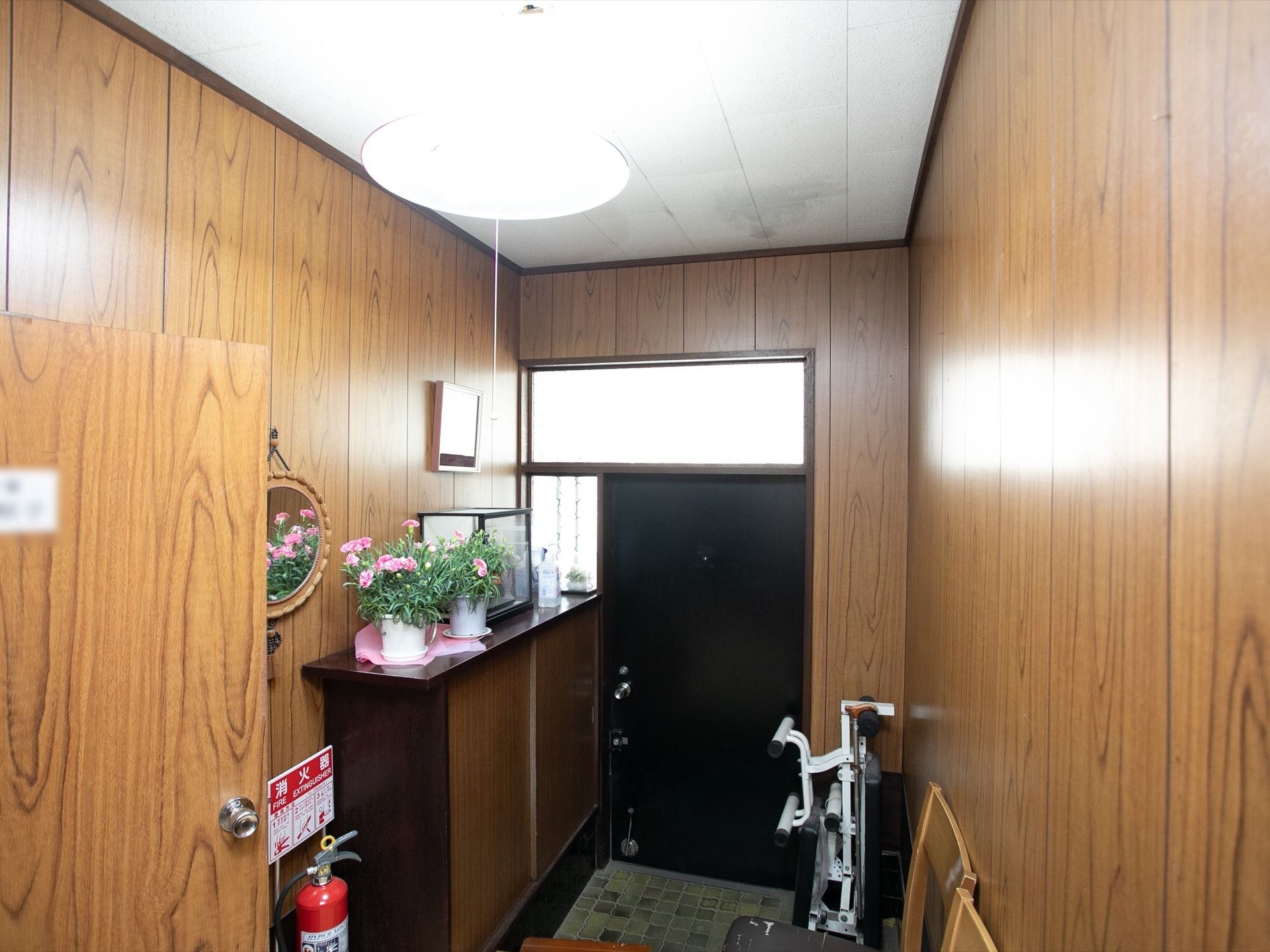 茶色の木目を基調とした戸建て住宅風の玄関。下駄箱の上には、さまざまな置物や手指の消毒スプレーなどが並べられ、近くに椅子が置かれている。