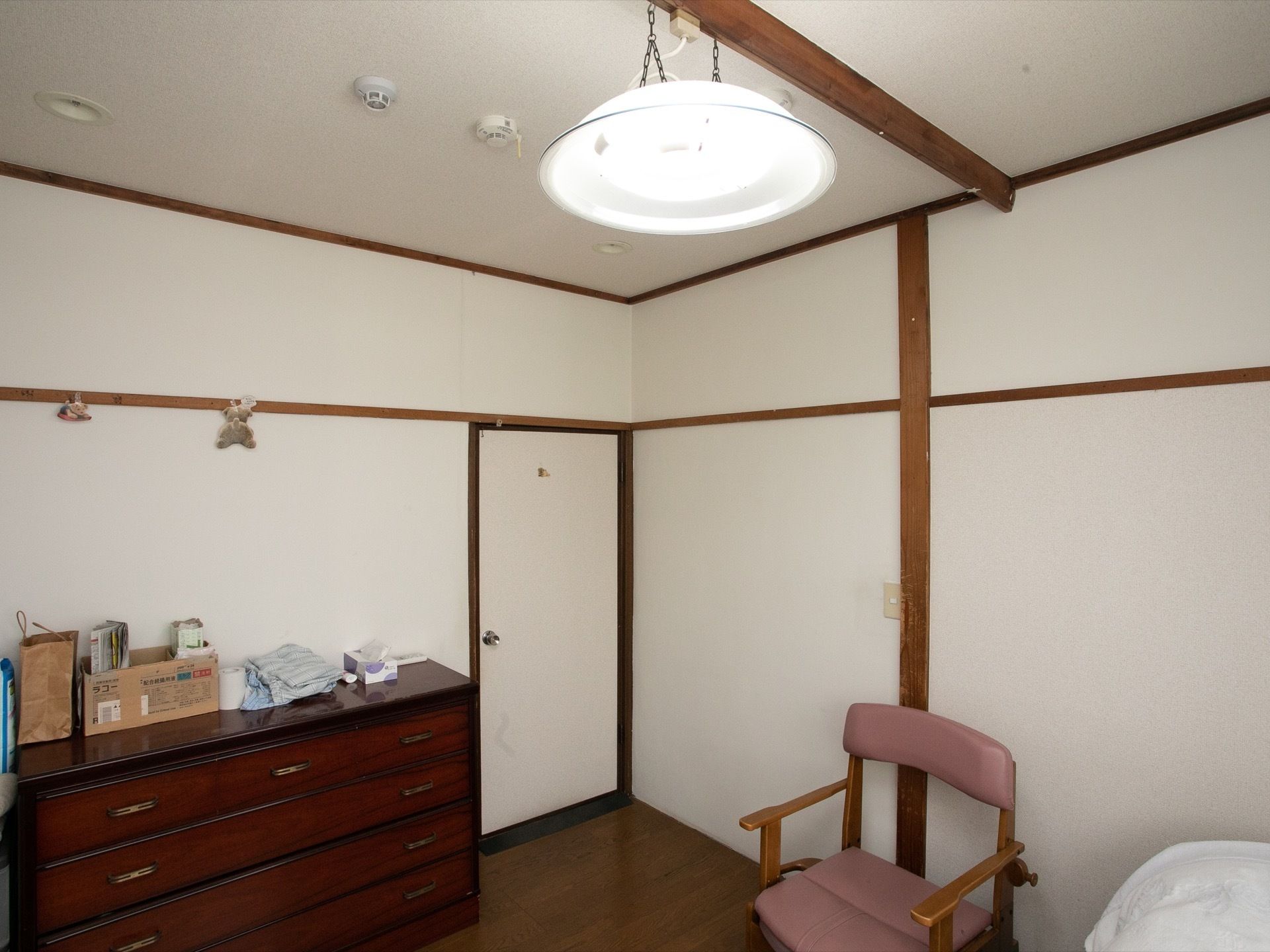 ホーム内の居室。濃い茶色の収納用のタンスや、茶色の枠にピンクの布張りの椅子、照明などが設置されている。