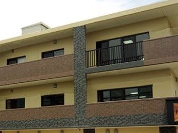 薄い黄色の外壁の建物。２階以上の部分にはベランダがあり、転落防止のためブラウンとグレーを基調とした壁が設置されている。
