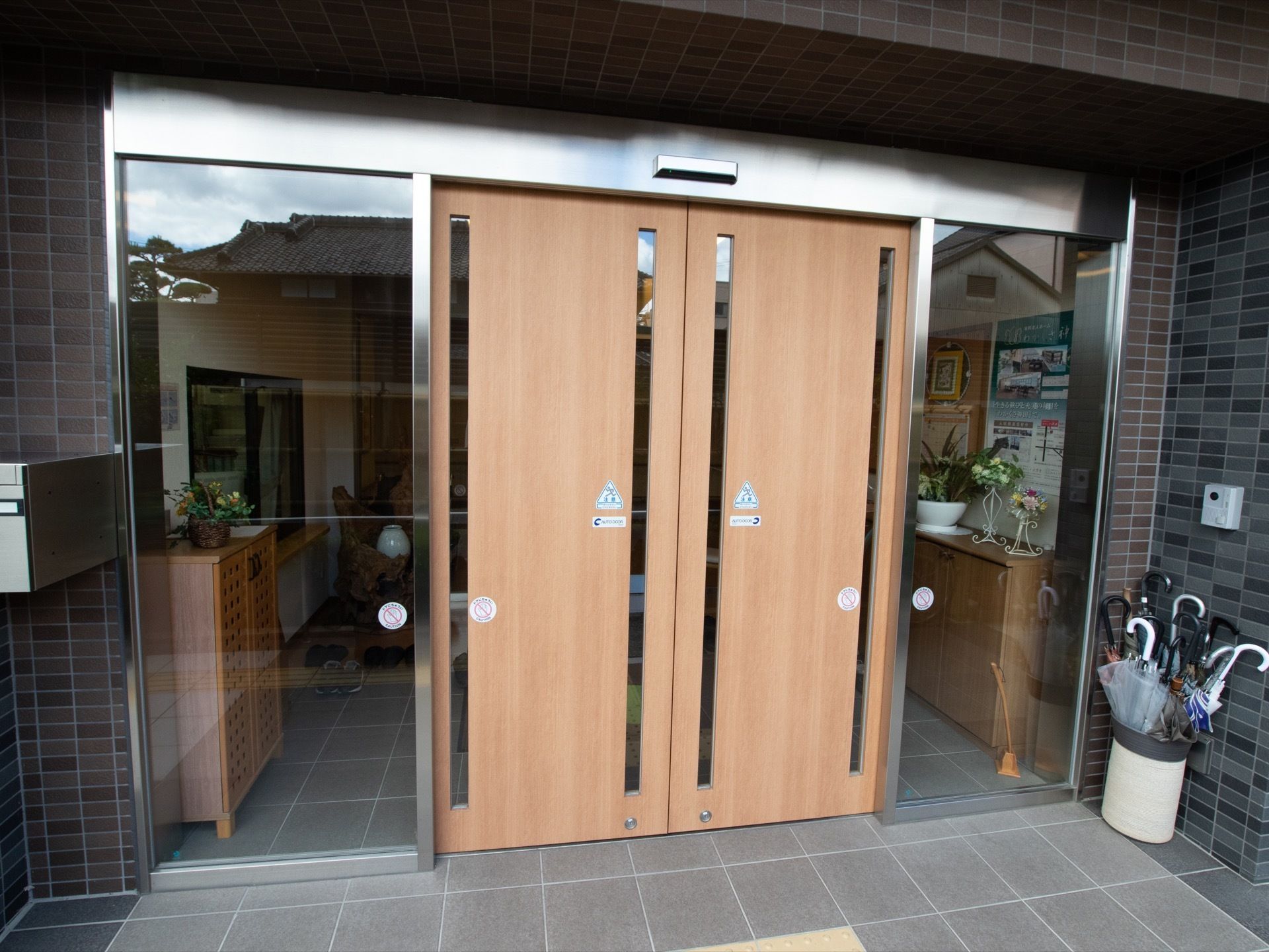 木目調の自動ドアを取り入れた玄関である。両側はガラス張りになっている。壁際の角に傘立てを置き、反対側の壁には郵便受けを取付けている。