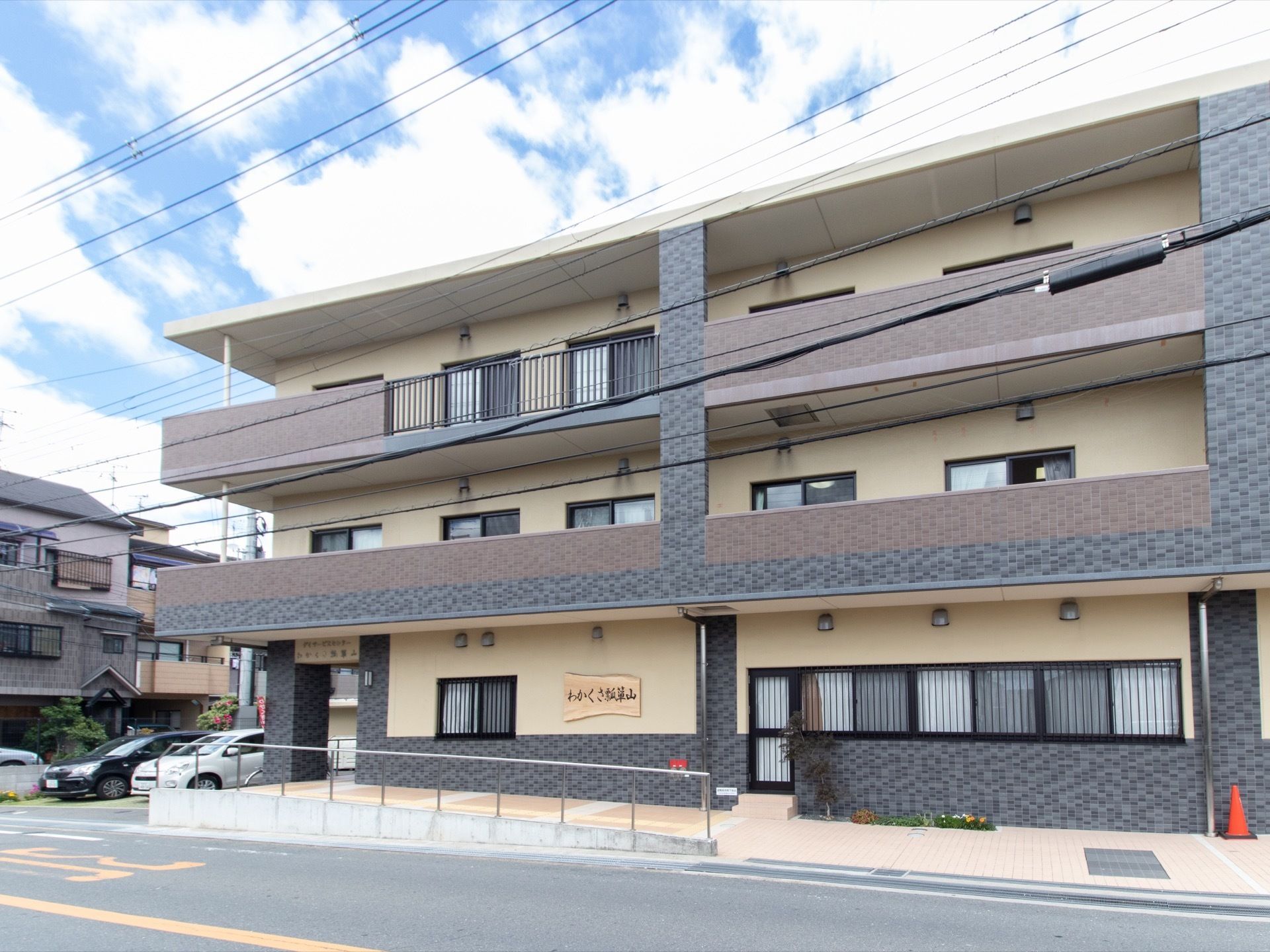 ベージュとブラウン、グレーの色合いの３階建ての外観である。１階の外壁に木造りの表札を掲げている。建物横にスロープを設けている。