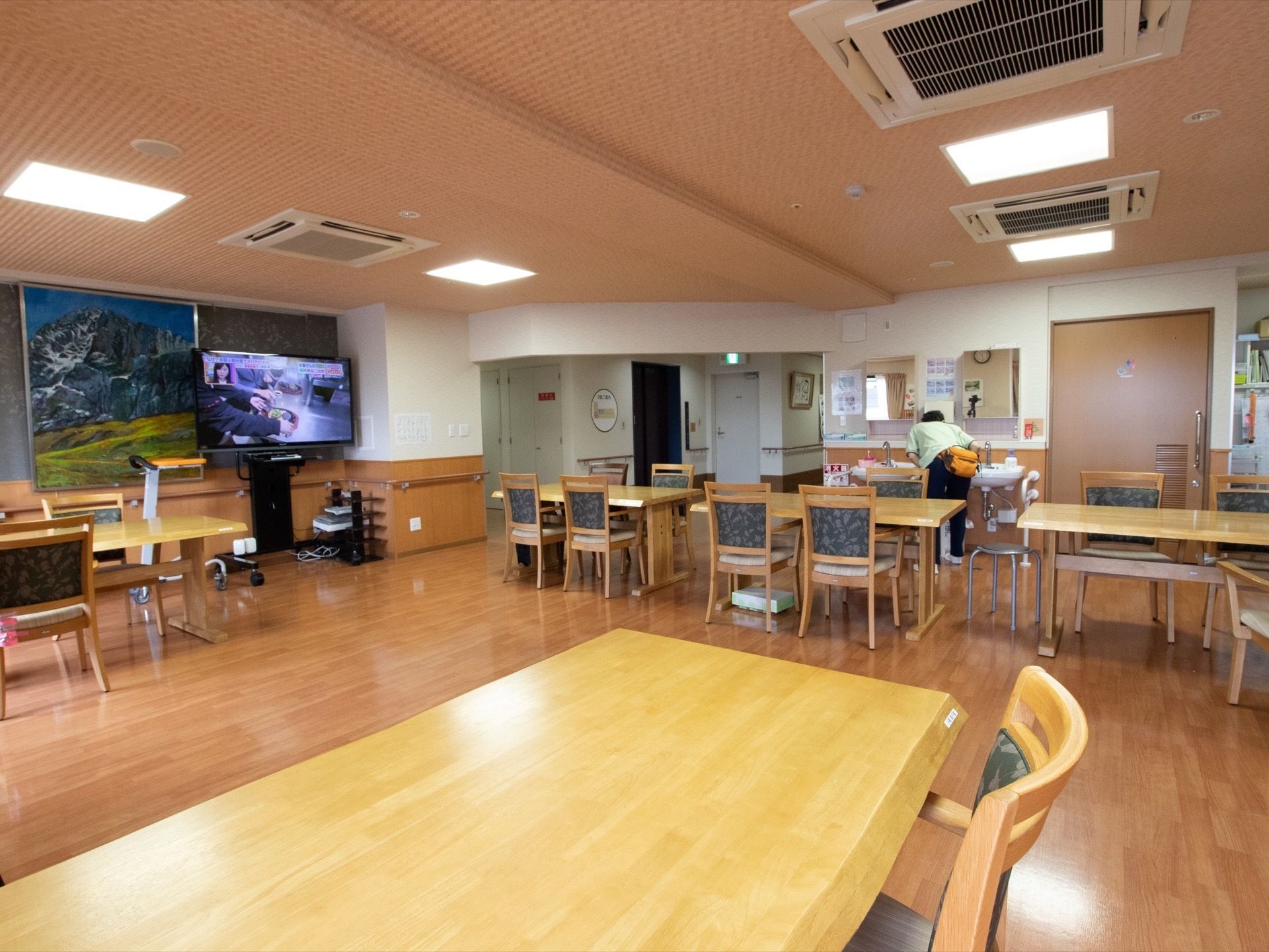4人掛けテーブルと椅子が並び、大きな壁掛け絵画の前にテレビが設置された広い食堂