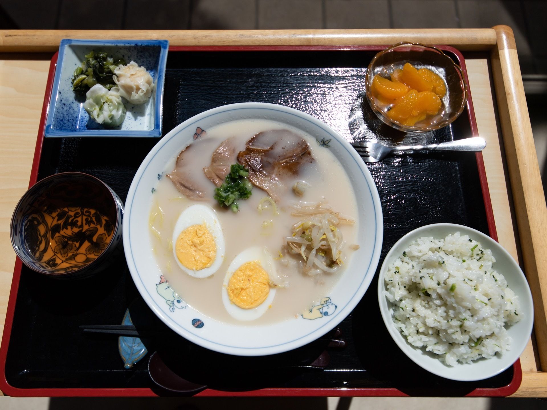 黒のトレーの中央にメインのラーメン、横に混ぜご飯を配置している。上部に小さな器の添え物とフルーツの小鉢を添えている。