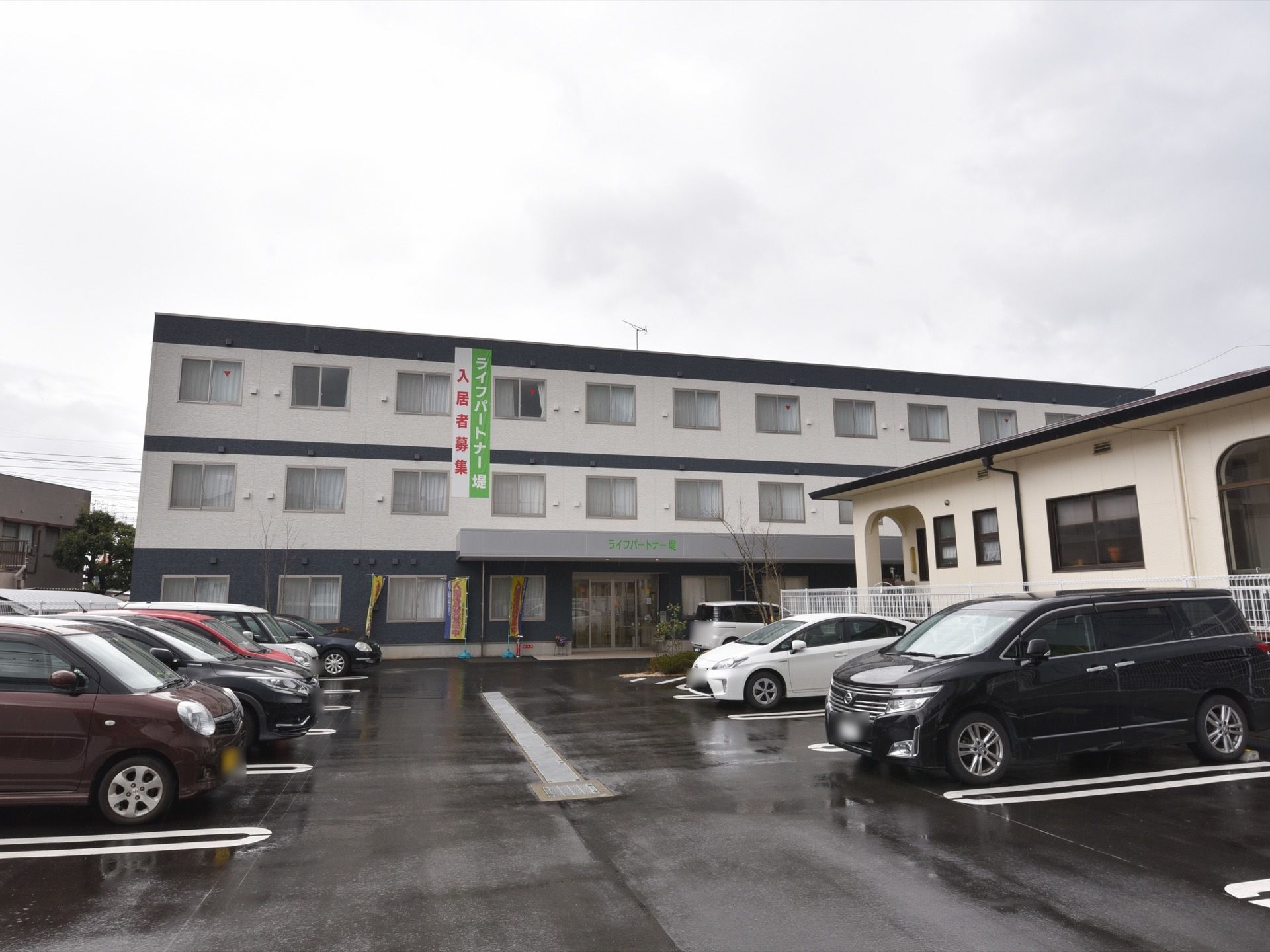 モノクロに塗装された３階建ての施設。玄関前には駐車スペースになっている。入居者募集の垂幕や幟が設置されている。