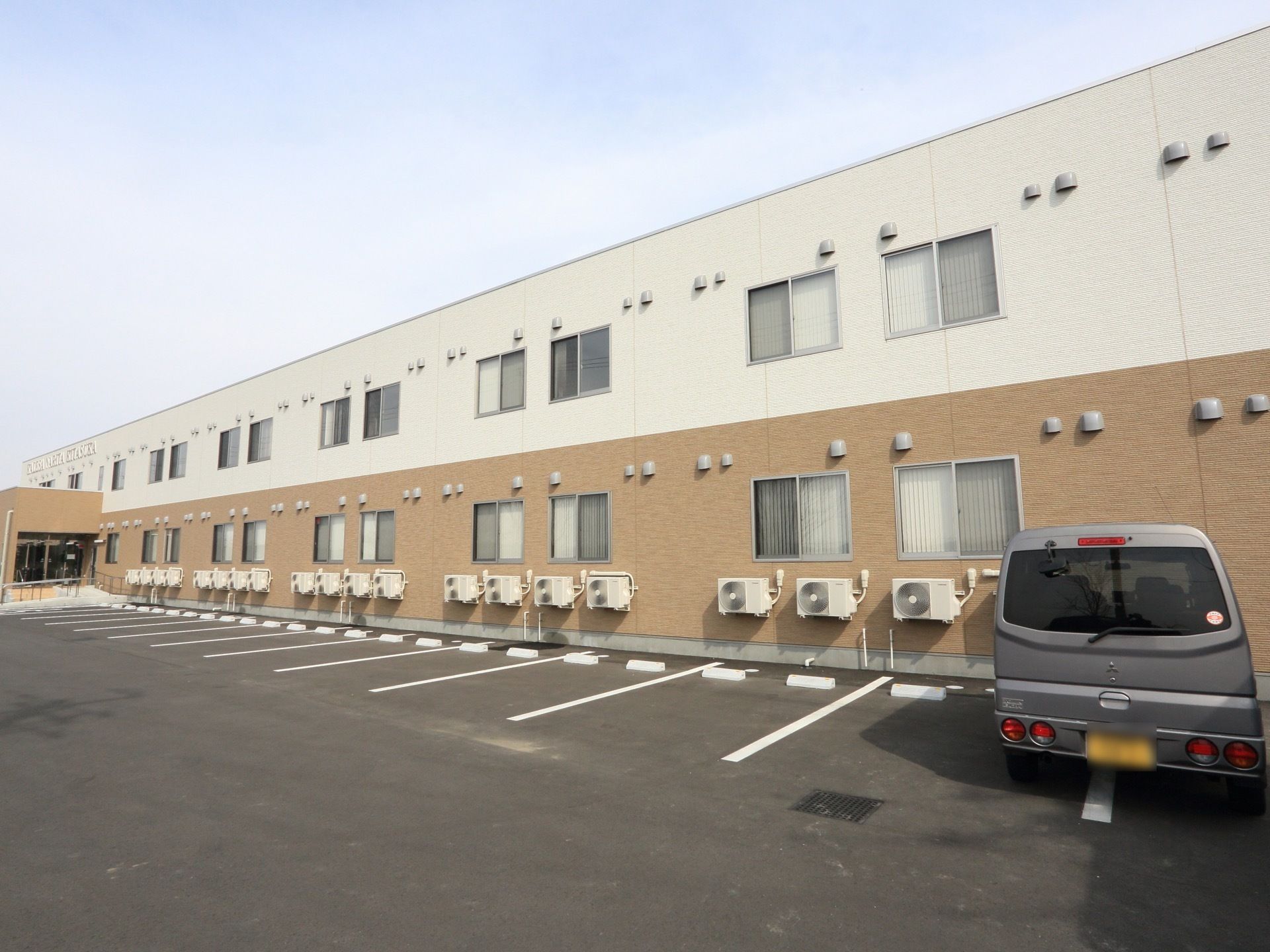 ２階建ての建物で、外壁が２色になっている。１階部分にすべての室外機を設置している。施設の前には広い駐車場がある。
