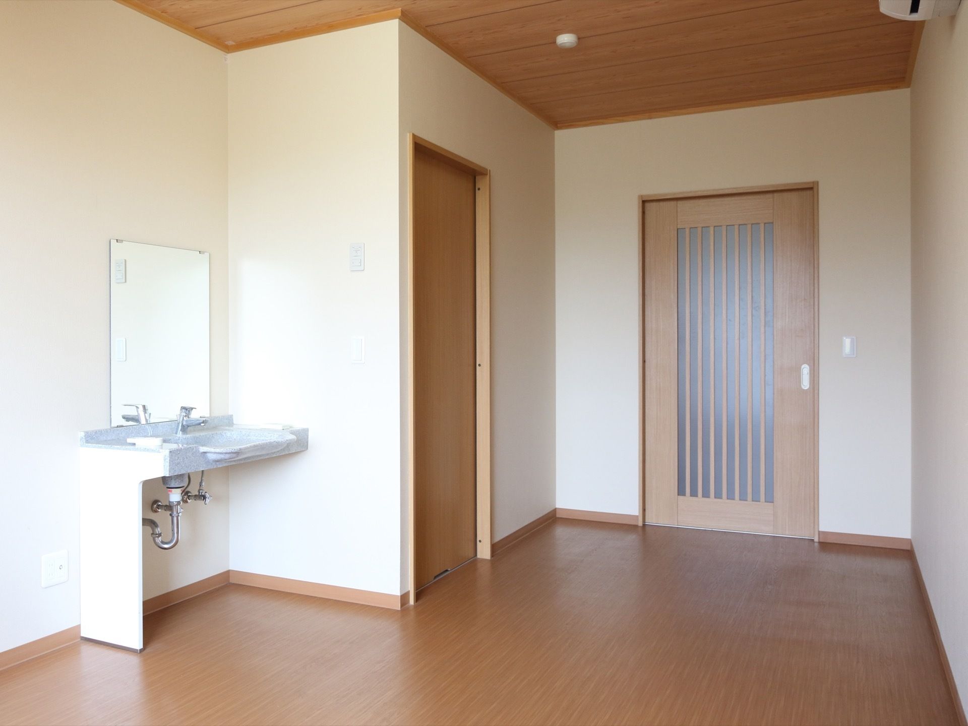居室は、木目調の床と天井、白い壁といったシンプルな内装となっている。左側には、簡易洗面台が設置されている。