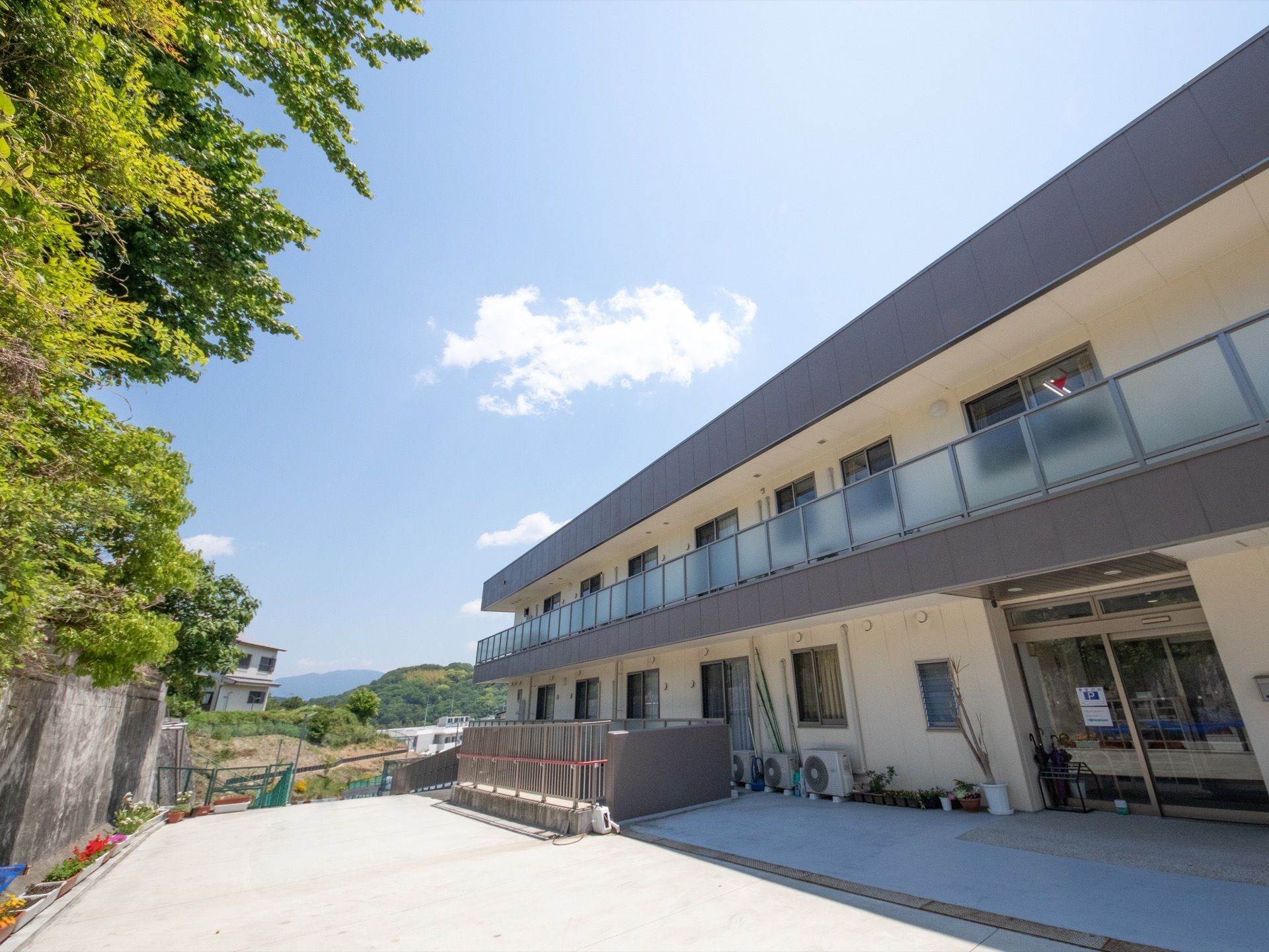 細長くクリーム色の施設の建物が伸びており、隣の境界との間には広いスペースの通路がある。緑の樹木と青い空が映える。