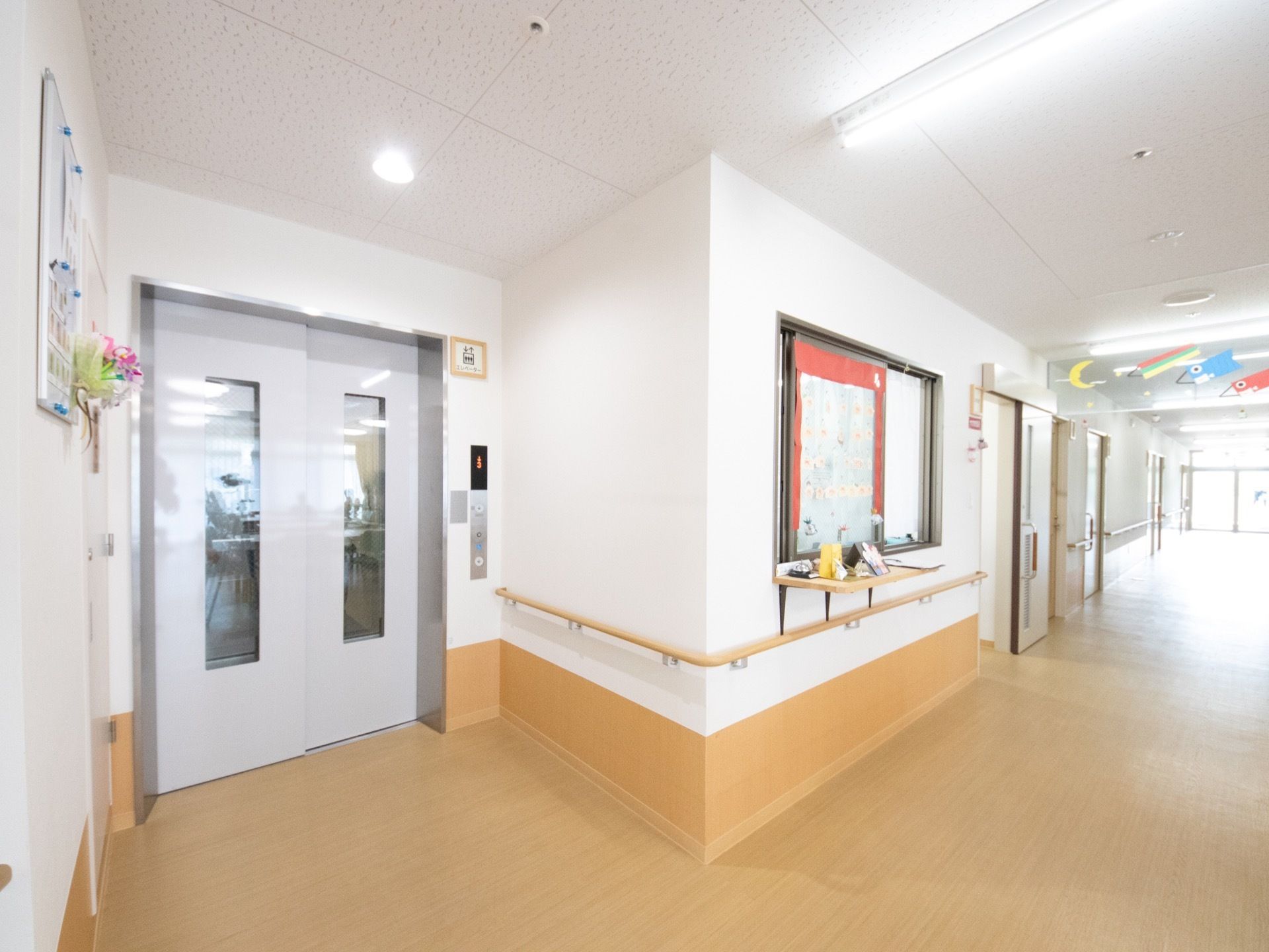 広いエレベータがあり、廊下の壁には手すりが備えられている。お花や季節を感じられる折り紙での装飾が施されている。