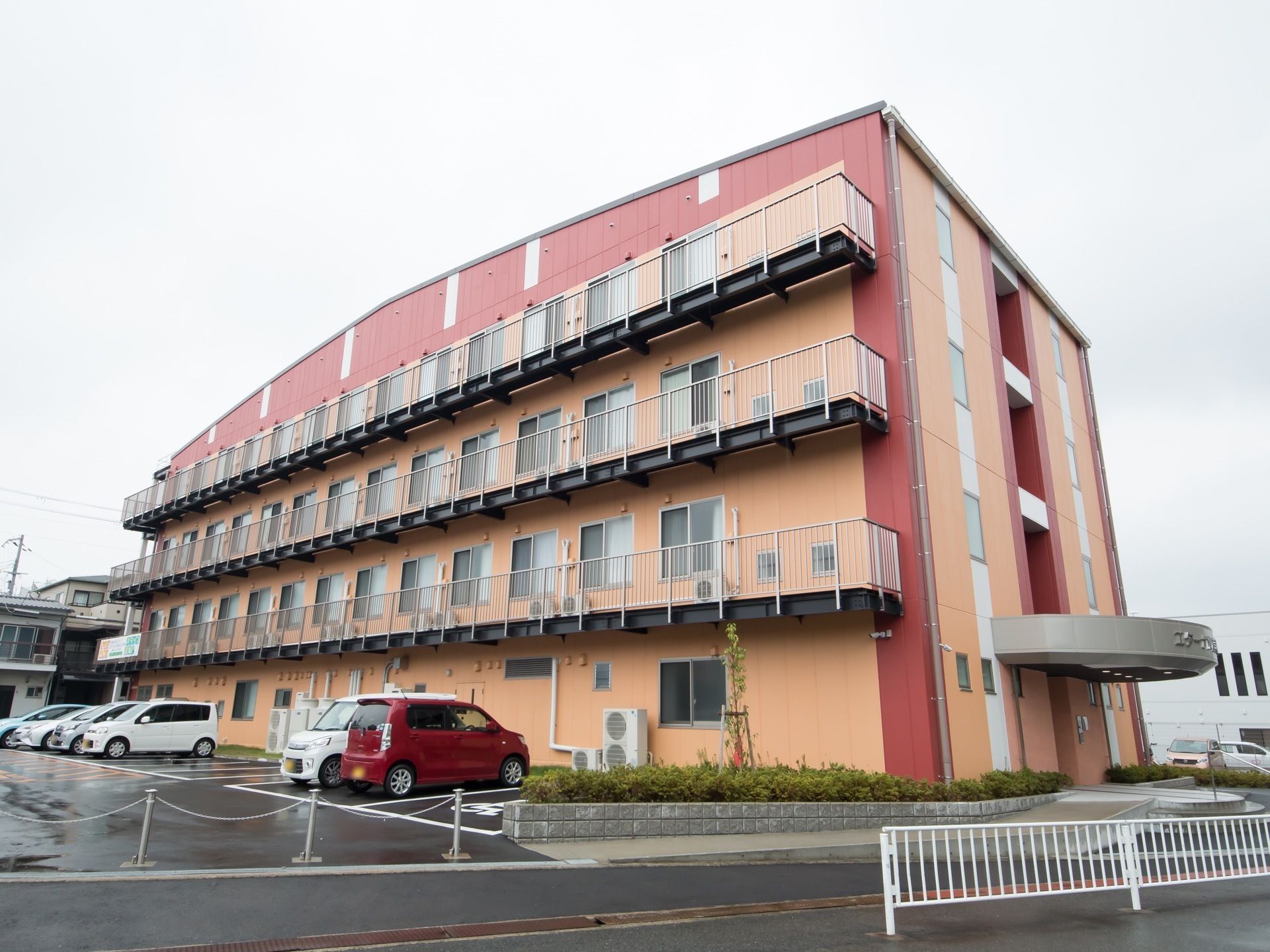 大きな施設の壁は、赤とオレンジでカラフルに塗装されている。施設には広い駐車場が併設され、複数の車が停まっている。