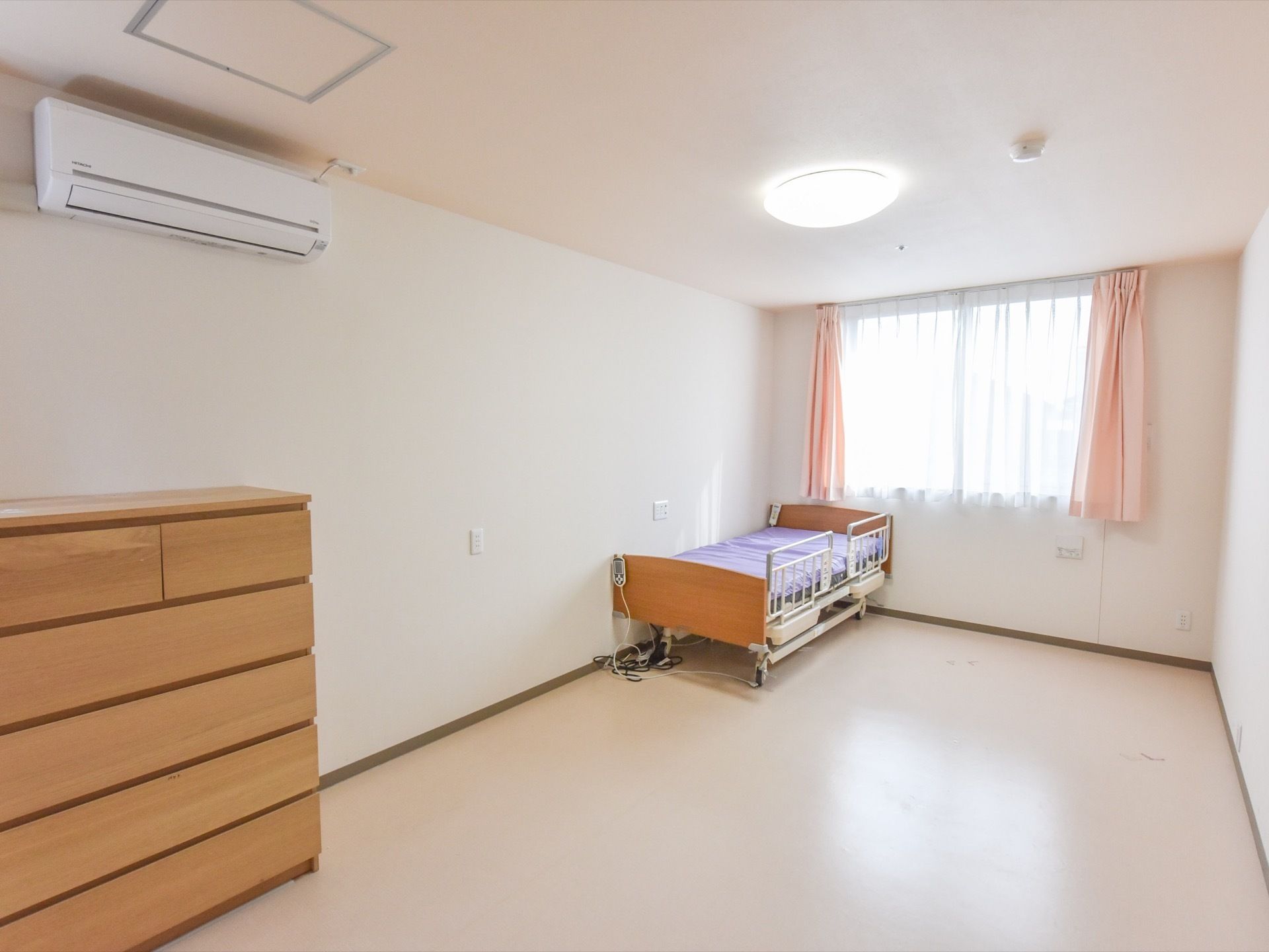 居室にはローチェストがあり、介護用ベッドが置かれている。また壁には窓があり、カーテンが取り付けられている。