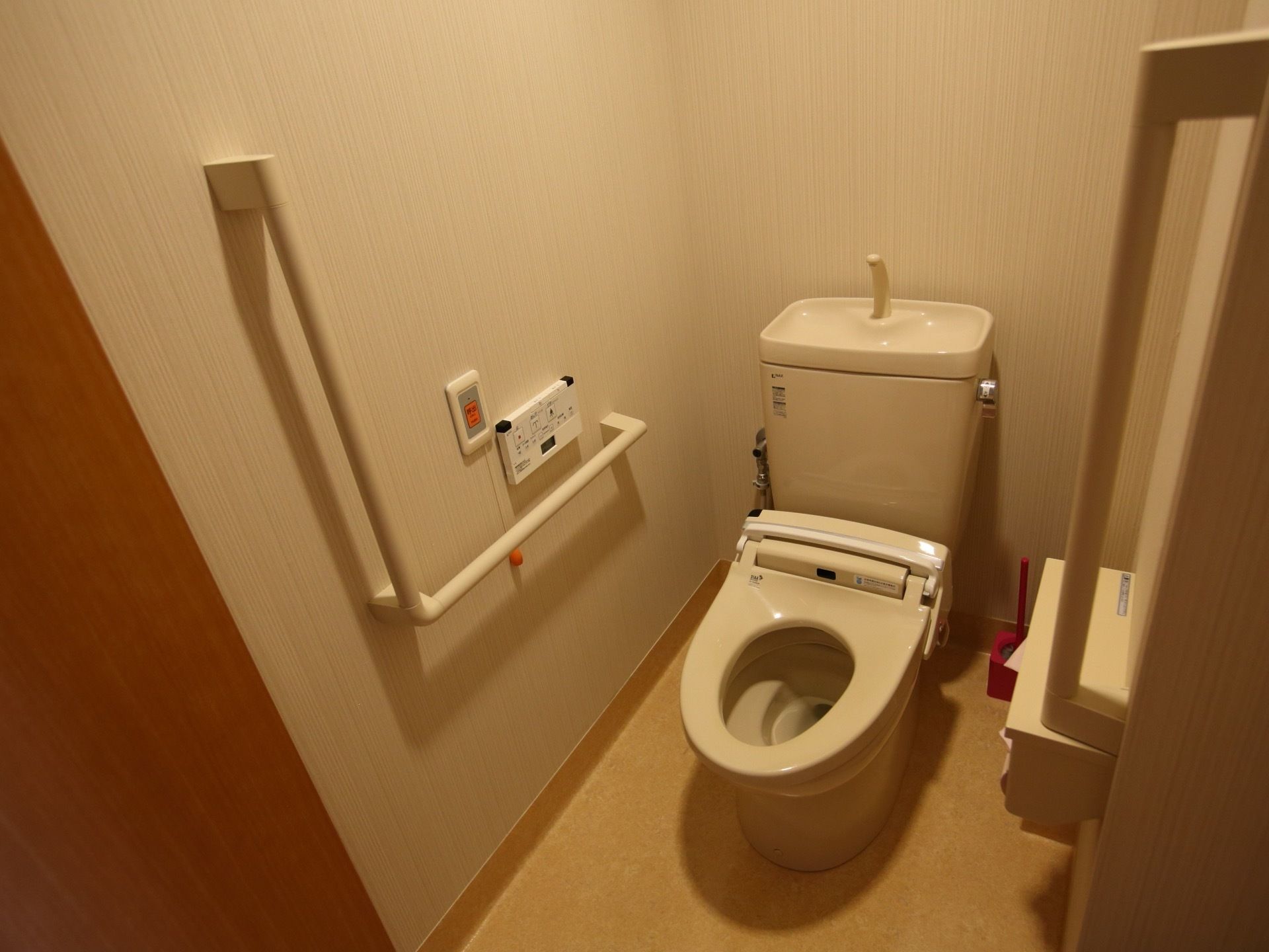 壁にＬ字の手すりが付いた洋式トイレがある。手すりのそばには操作パネルと緊急通報ボタンが設置されている。