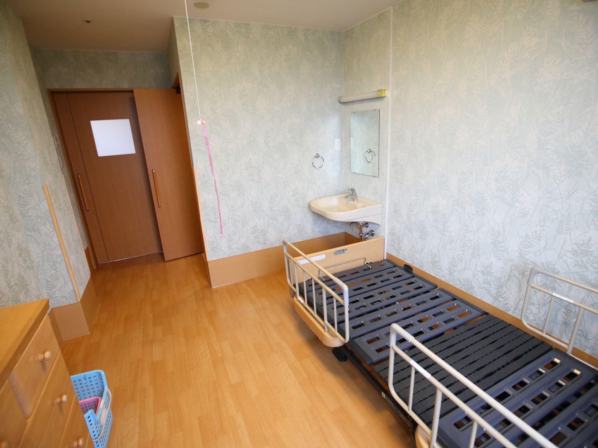 フローリングの居室に、電動ベッドと木製のチェストが置かれている。ベッドの足元には洗面台も設置されている。