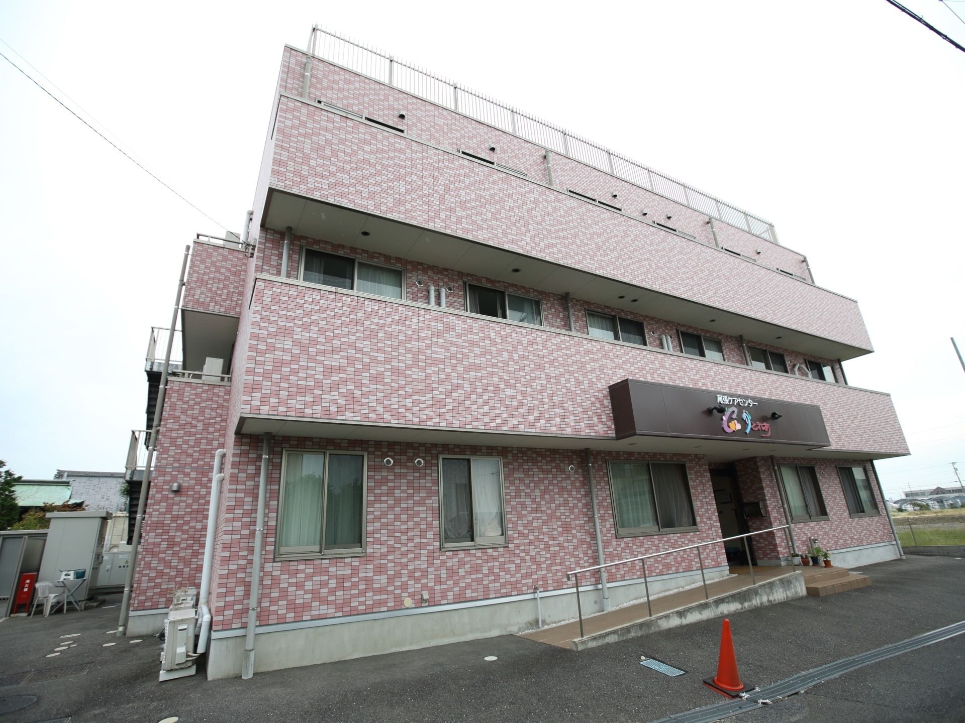 ３階建ての住宅があり、外壁は白や淡いピンクのタイルが貼られている。敷地は舗装され、玄関の前には階段と手すり付きのスロープがある。