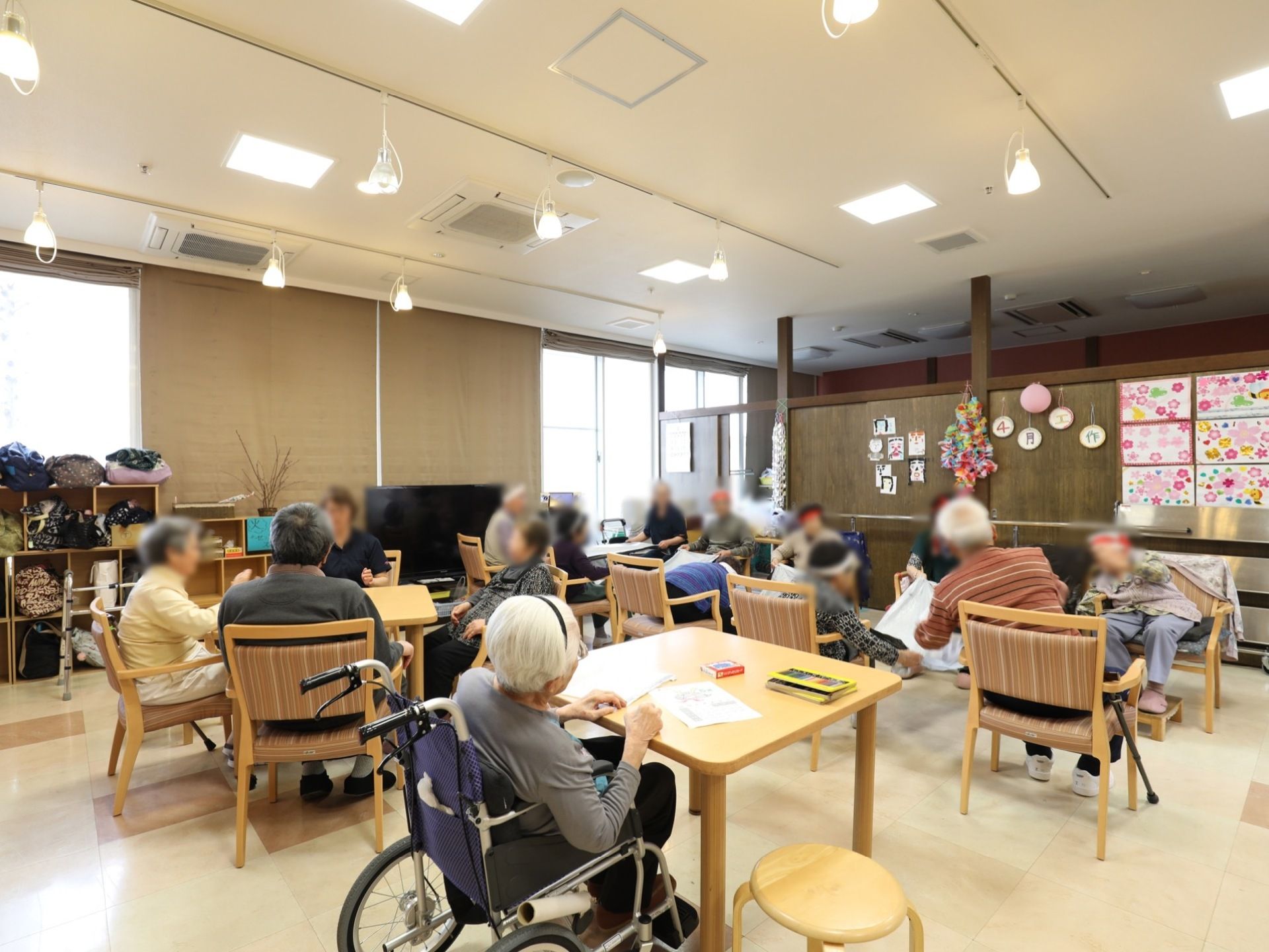 いくつものテーブルが置かれ、入居者様が思い思いに過ごしている。窓がわにはロッカーやテレビが置かれている。