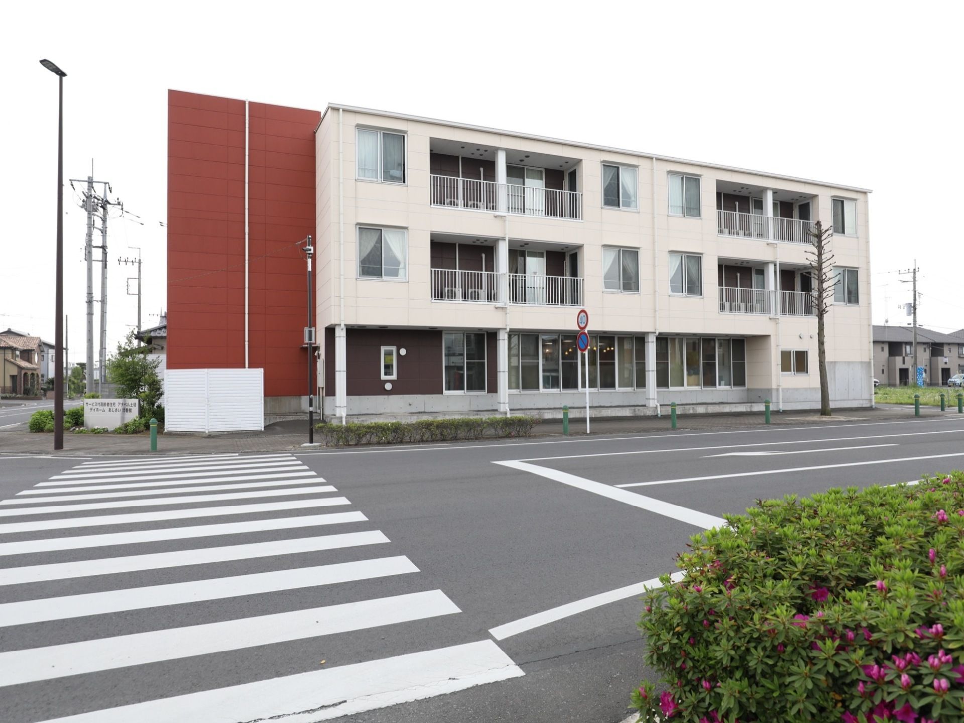 3階建ての建物で、白とダークブラウンの外壁である。左側に赤色の外壁の部分がある。敷地の角に、施設名の書かれた看板が立っている。