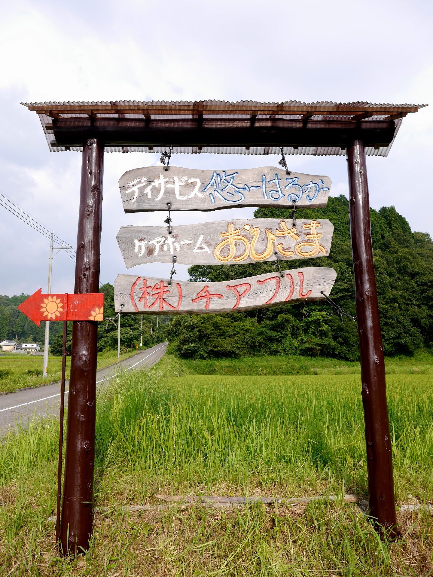 田んぼのあぜ道に設置されたトタンの屋根が付いた手作りの看板。赤い矢印もついている。