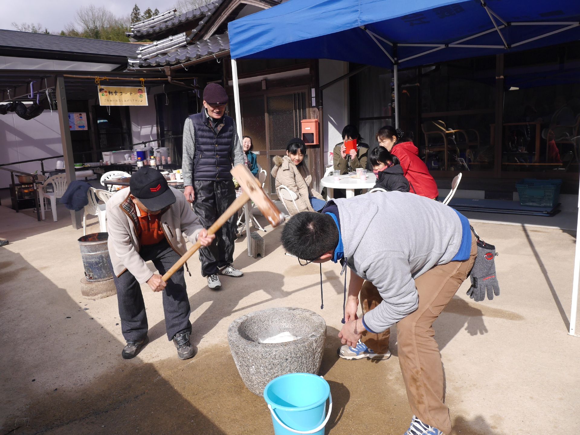 日本家屋の庭で高齢者が餅つきをしている。