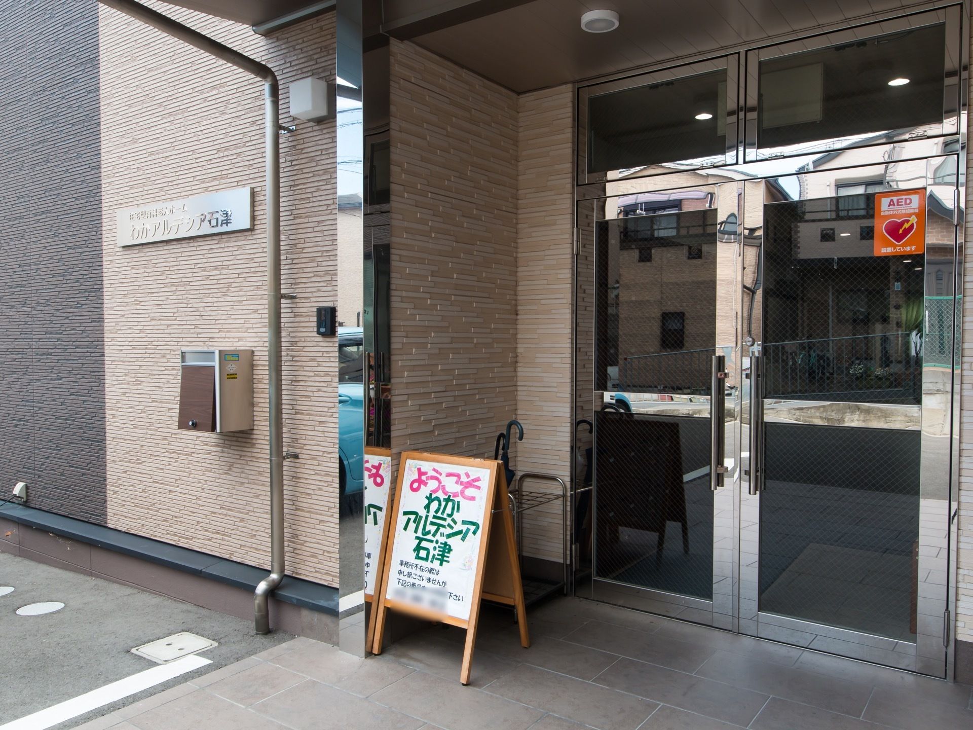 玄関はレバーを引く開き戸になっている。扉の右上に、AED設置のステッカーが貼られている。施設名称を表示したウェルカムボードが置かれている。