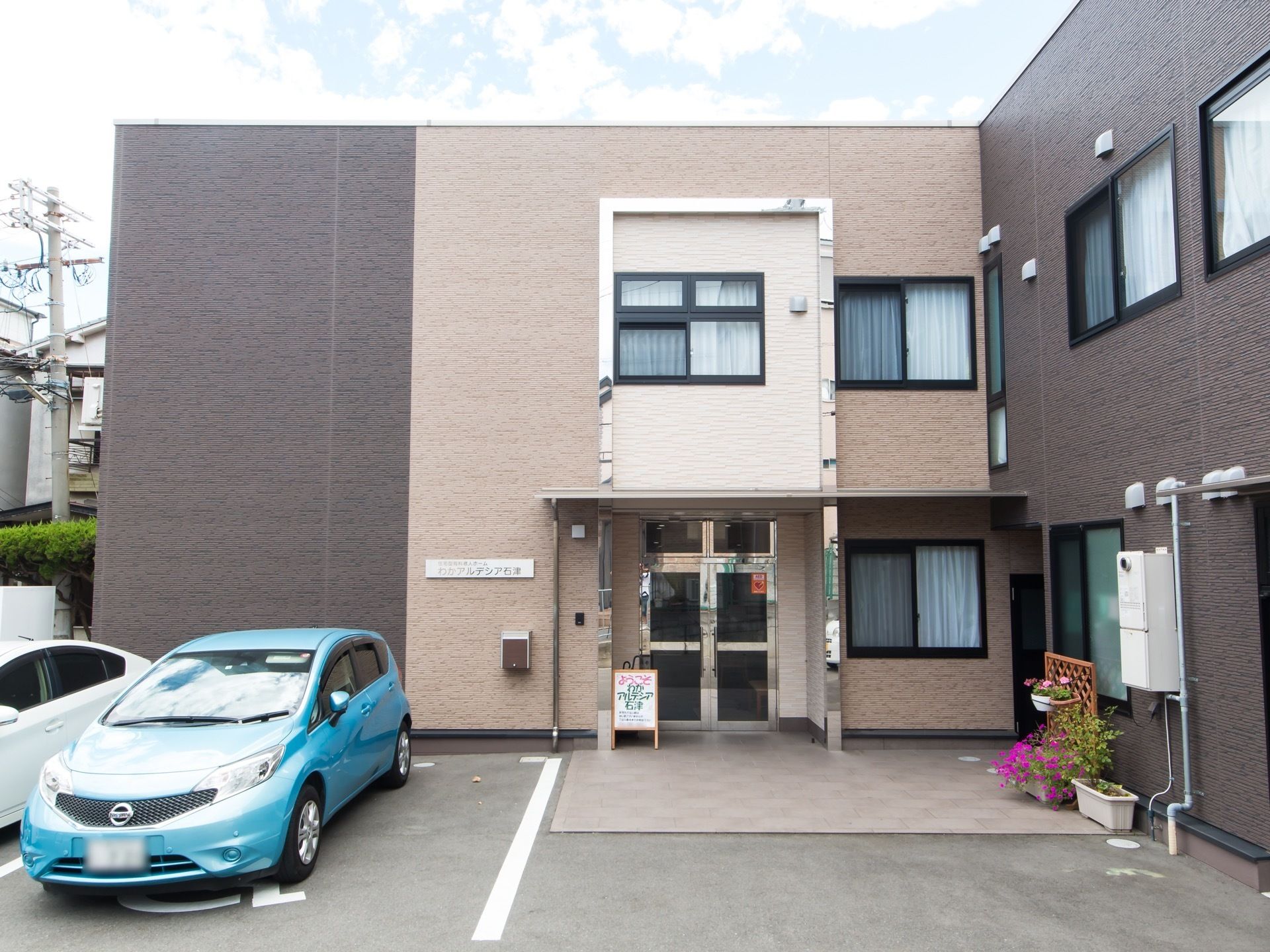 茶色系統の色を基調とした外壁の２階建て施設。玄関の左側に車椅子のマークが描かれた駐車スペースがある。