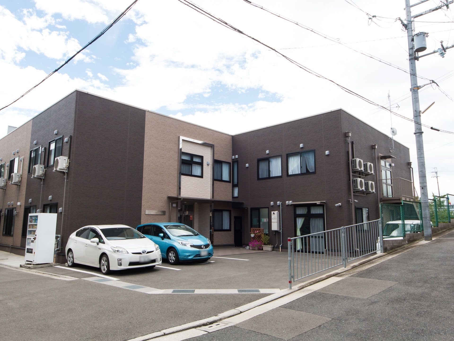 茶色系統の色を基調とした施設である。玄関の左側の敷地内に車２台分の駐車スペースがある。その横に飲料の自動販売機がある。