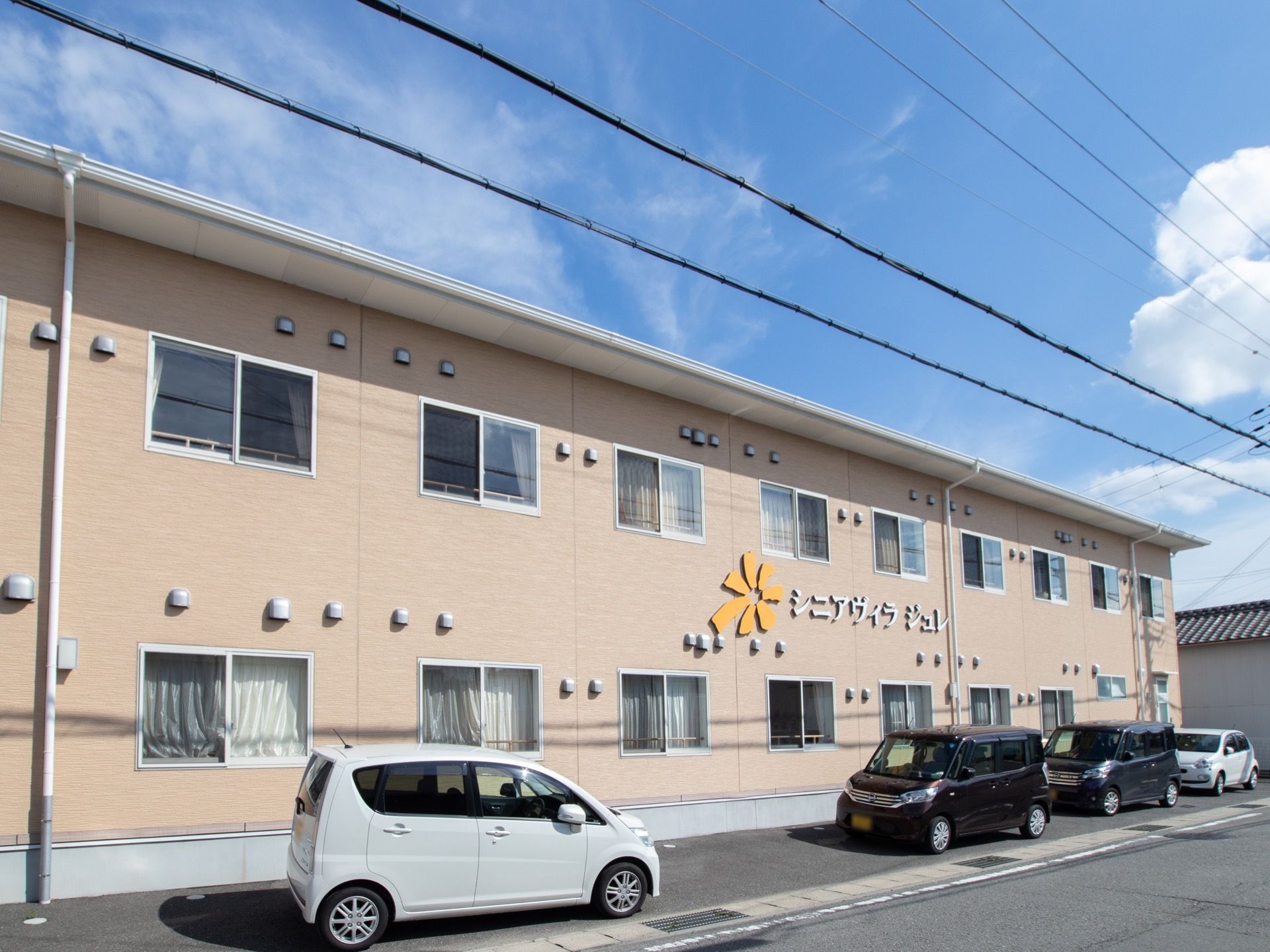 二階建ての横に長い建物で、壁にはオレンジ色の花の形のシンボルマークと、シルバーの施設名が大きく表示されている。