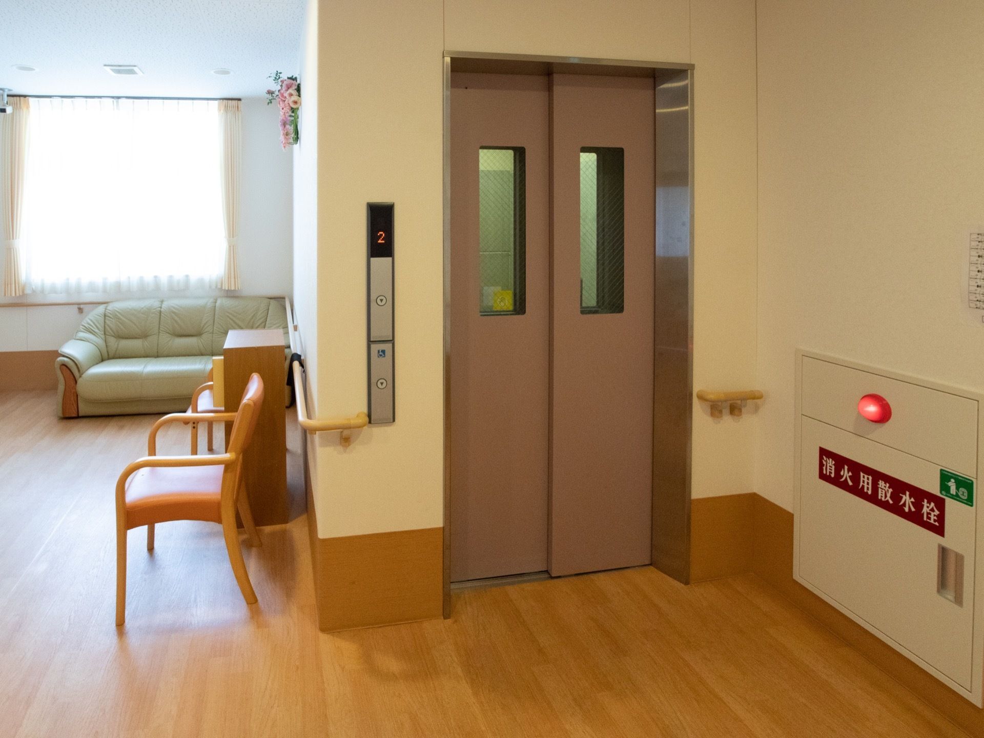 廊下の一角にエレベーターが設置されており、その前の壁に消火用撒水栓が備わっている。左側に披露スペースがあり、ソファや椅子が置かれている。