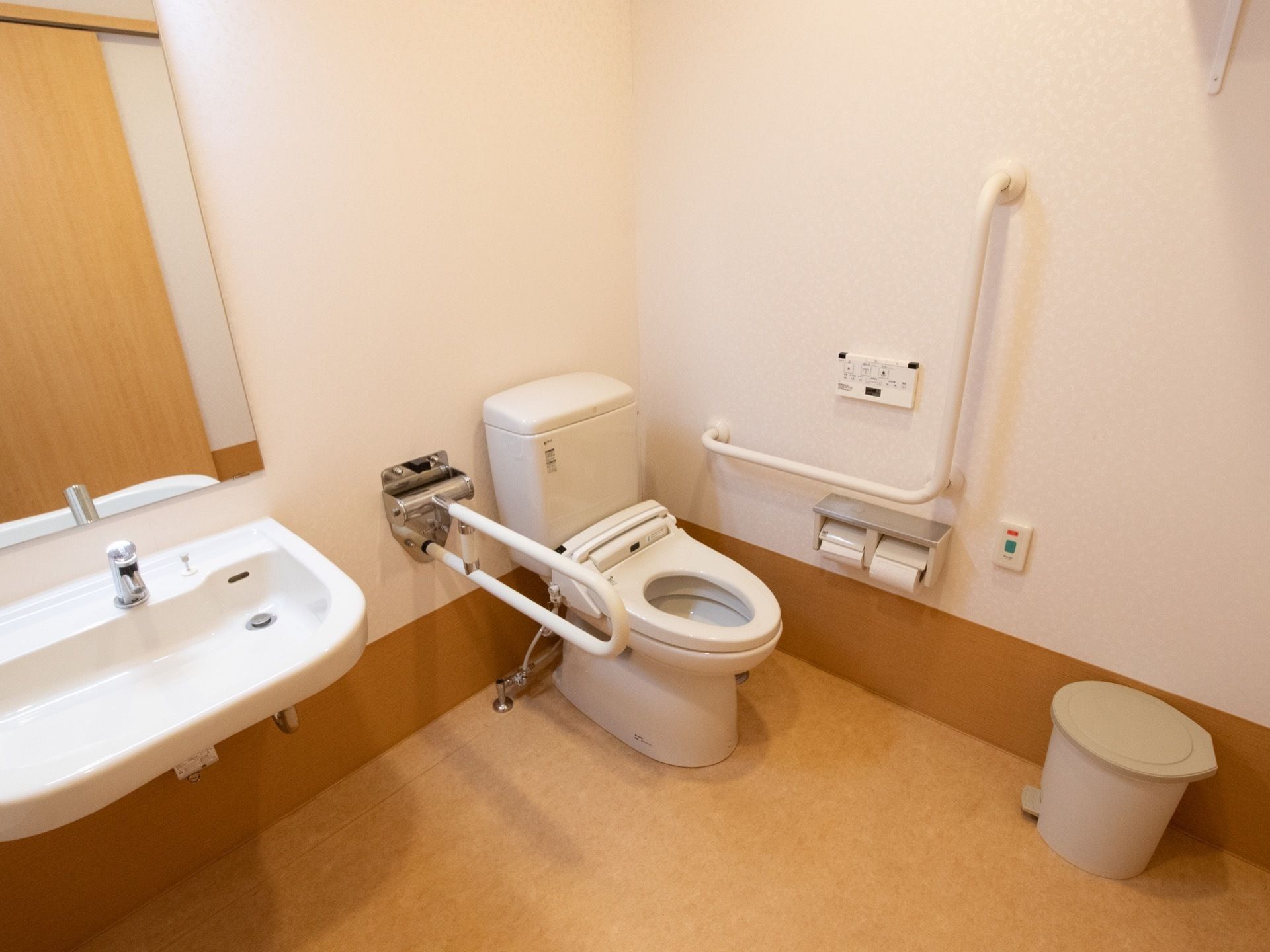 広いスペースのある個室内に、洗面台とトイレが設置されている。便器脇には可動式の手すりが備わっている。