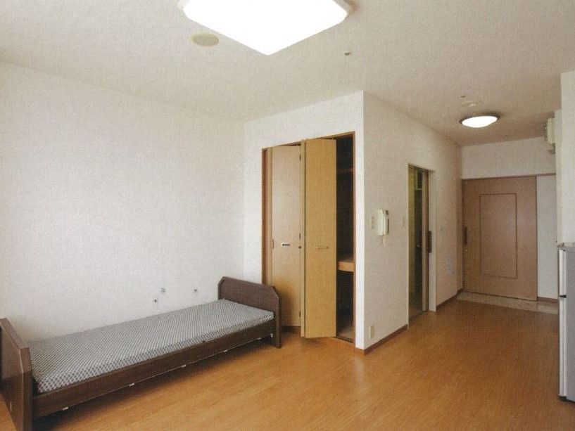 奥から見た部屋の様子。壁や天井は白色で、床は木目調のフローリングである。左側にベッドが設置されている。