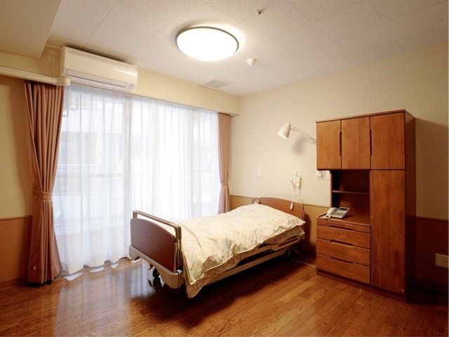 白い壁との木目調のフローリングが爽やかな居室だ。大きな窓が特徴的で明るい日差しが部屋に差し込んでいる。窓の側にベッドが置かれている。