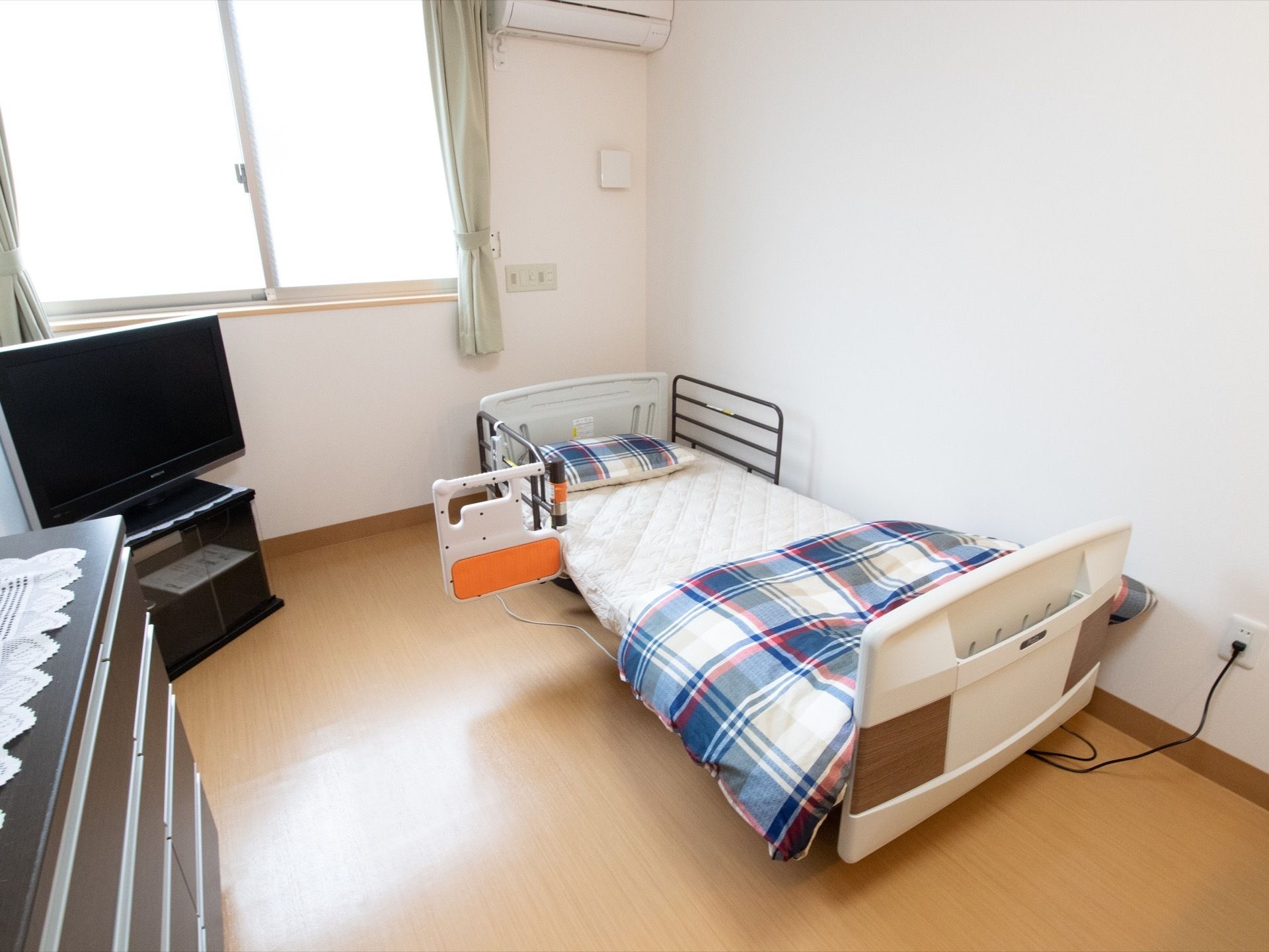 居室にはエアコンや窓が設置され明るい。補助器具付きの介護用ベッドやテレビ、整理タンスが置かれている。