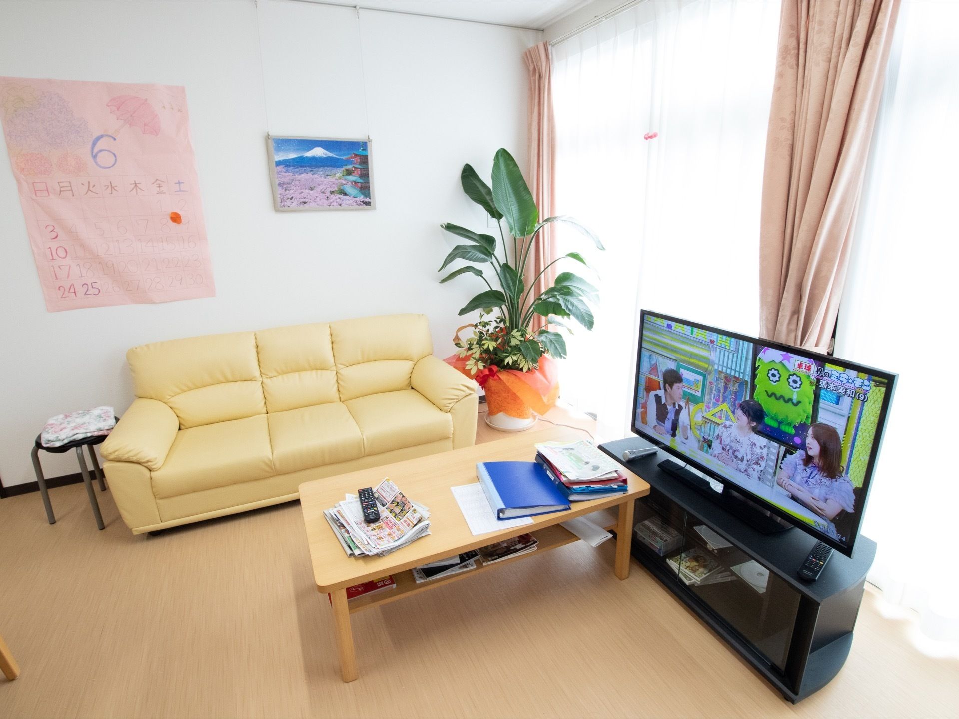 リビングの壁には手書きのイラスト入りカレンダーが貼られている。窓際には大きな観葉植物、ソファ、テレビが置かれている。
