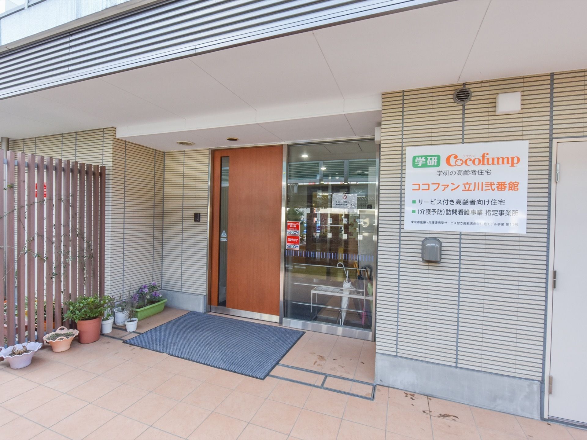 広々とした玄関前スペースである。横側の外壁に、施設名、施設種類などの看板を掲げている。出入口ドア前には、ブルーのマットを敷設している。
