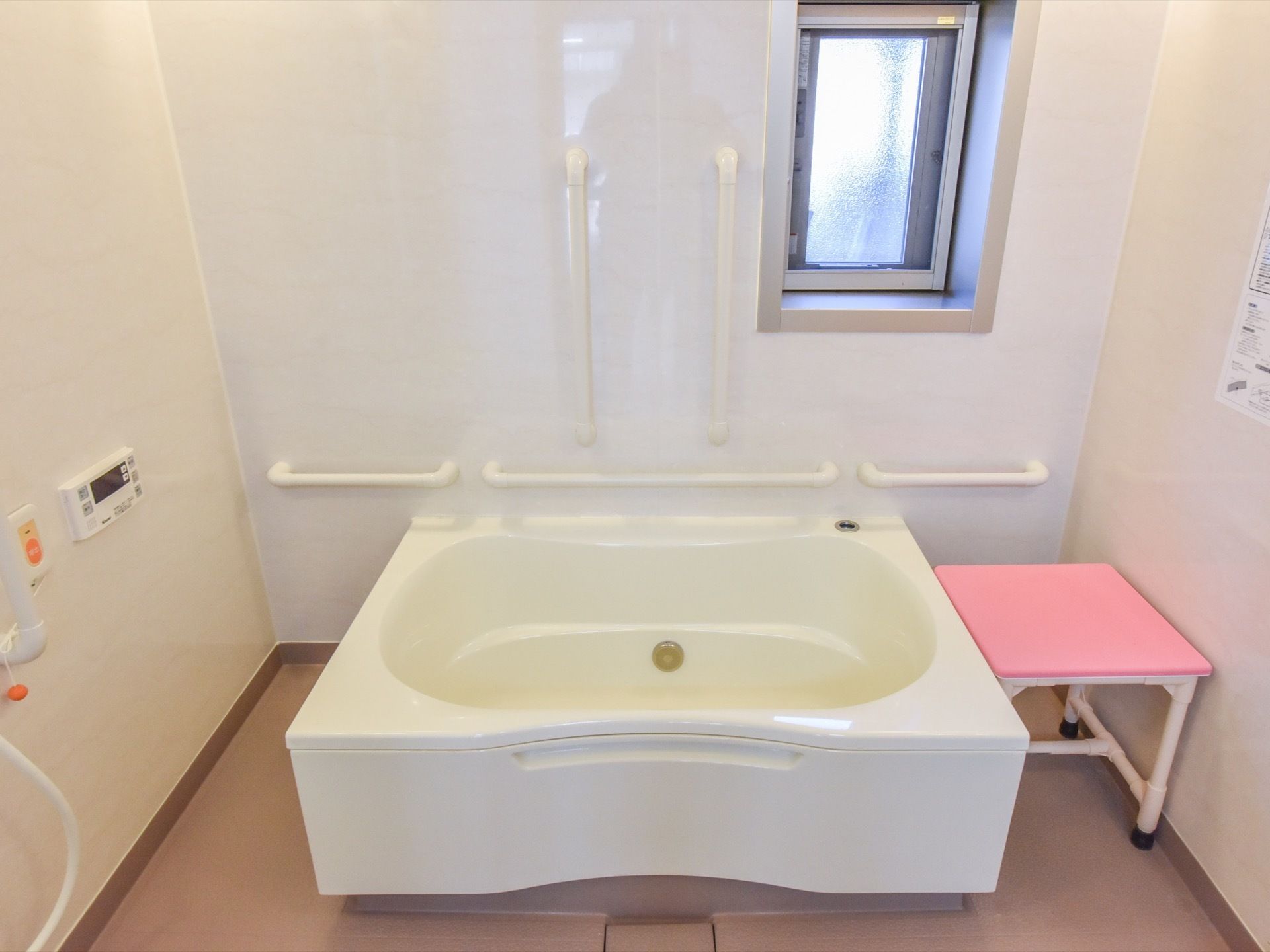 壁際に備えた浴槽の隣には、ピンク色の台がある。横側の壁際に縦に２本、横に３本の手すりを設置している。上部に長方形の小さな窓がある。