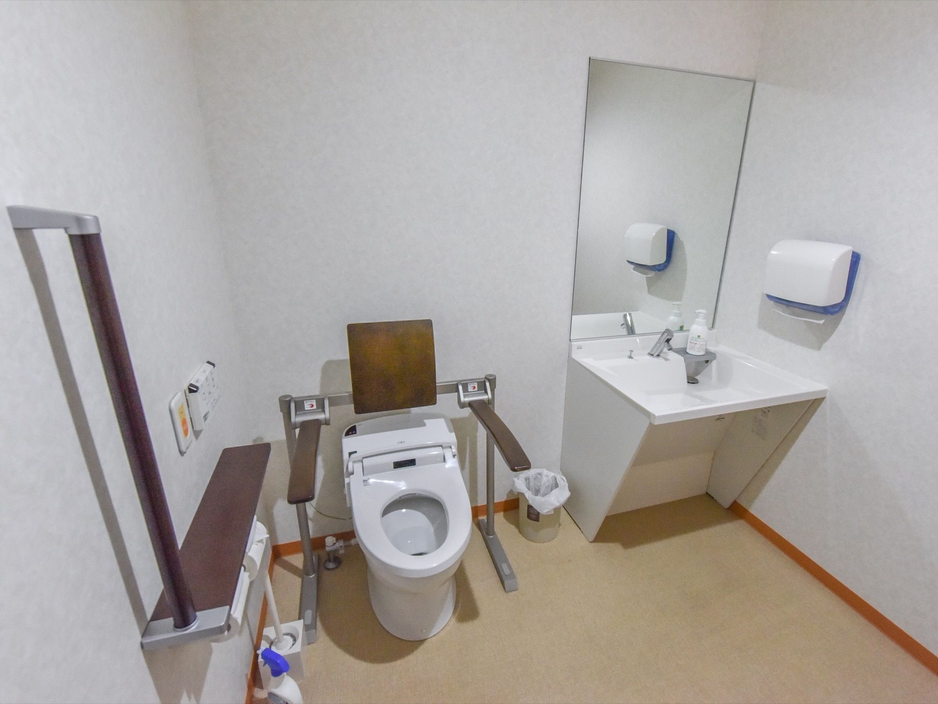 広々としたトイレスペースである。便器の隣に、白で統一した洗面台を設置している。便座の両側に手すり、後ろに背もたれを備えている。