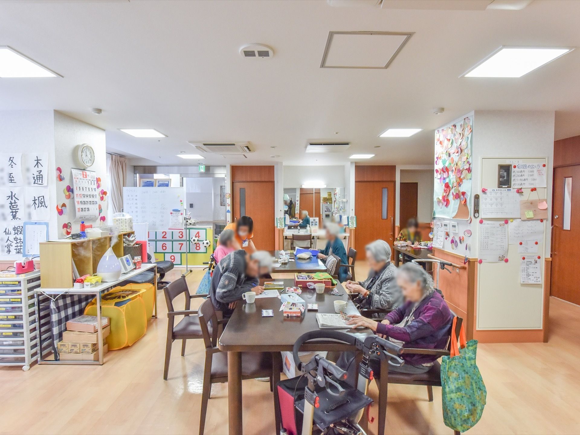 木目調のフローリングの共有スペースに、テーブルと椅子を並べている。壁には、入居者様の作品を貼り、カレンダーや手作りの花などを飾っている。