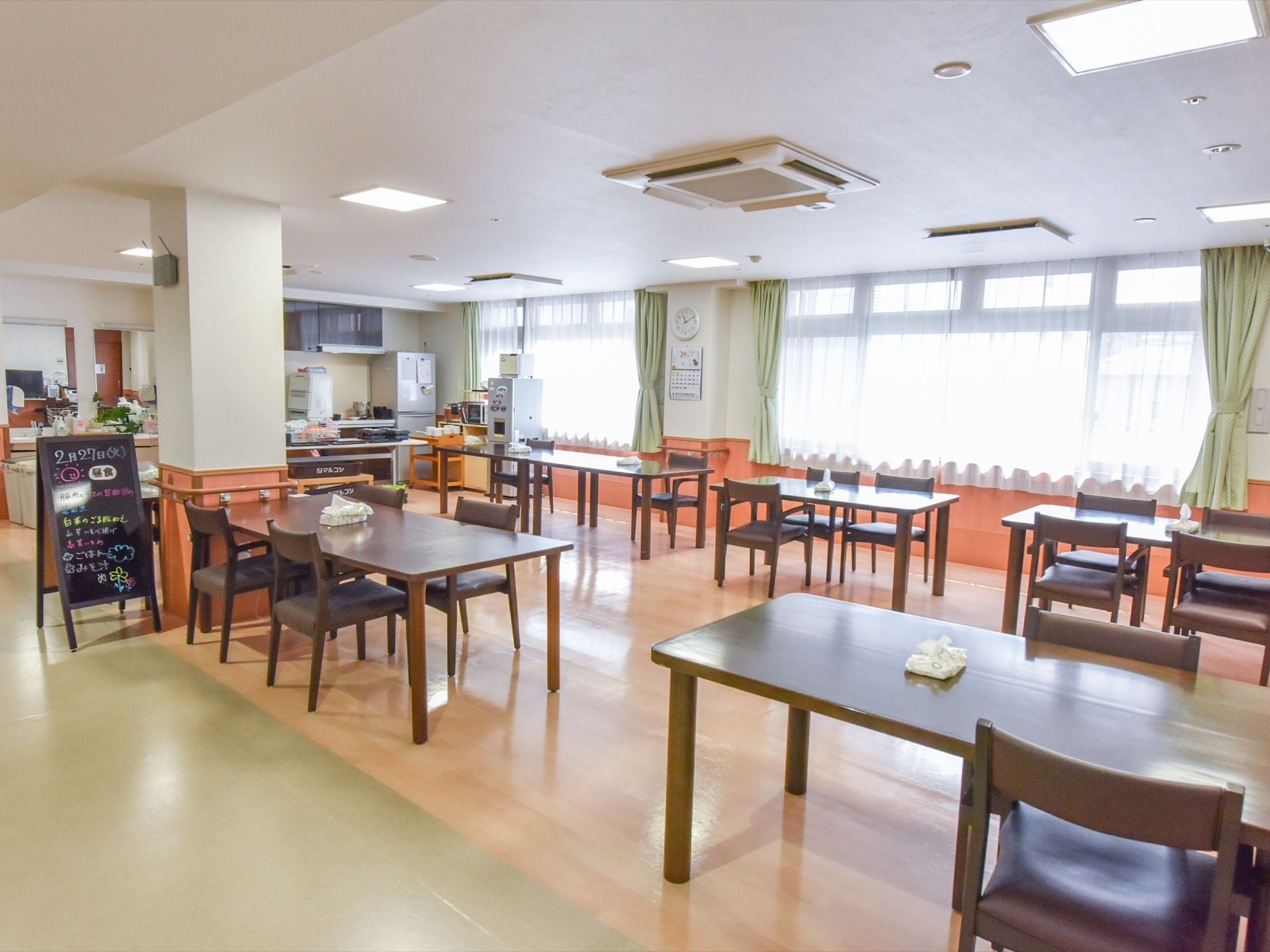 側面に窓が並び、明るい空間の食堂である。柱の隣に食事メニューを書いた黒面のボードを置いている。横側に厨房、洗面台スペースを設けている。