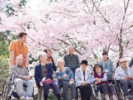桜の花の下で微笑んでいる高齢者と施設スタッフの様子。満開の花が咲いている
