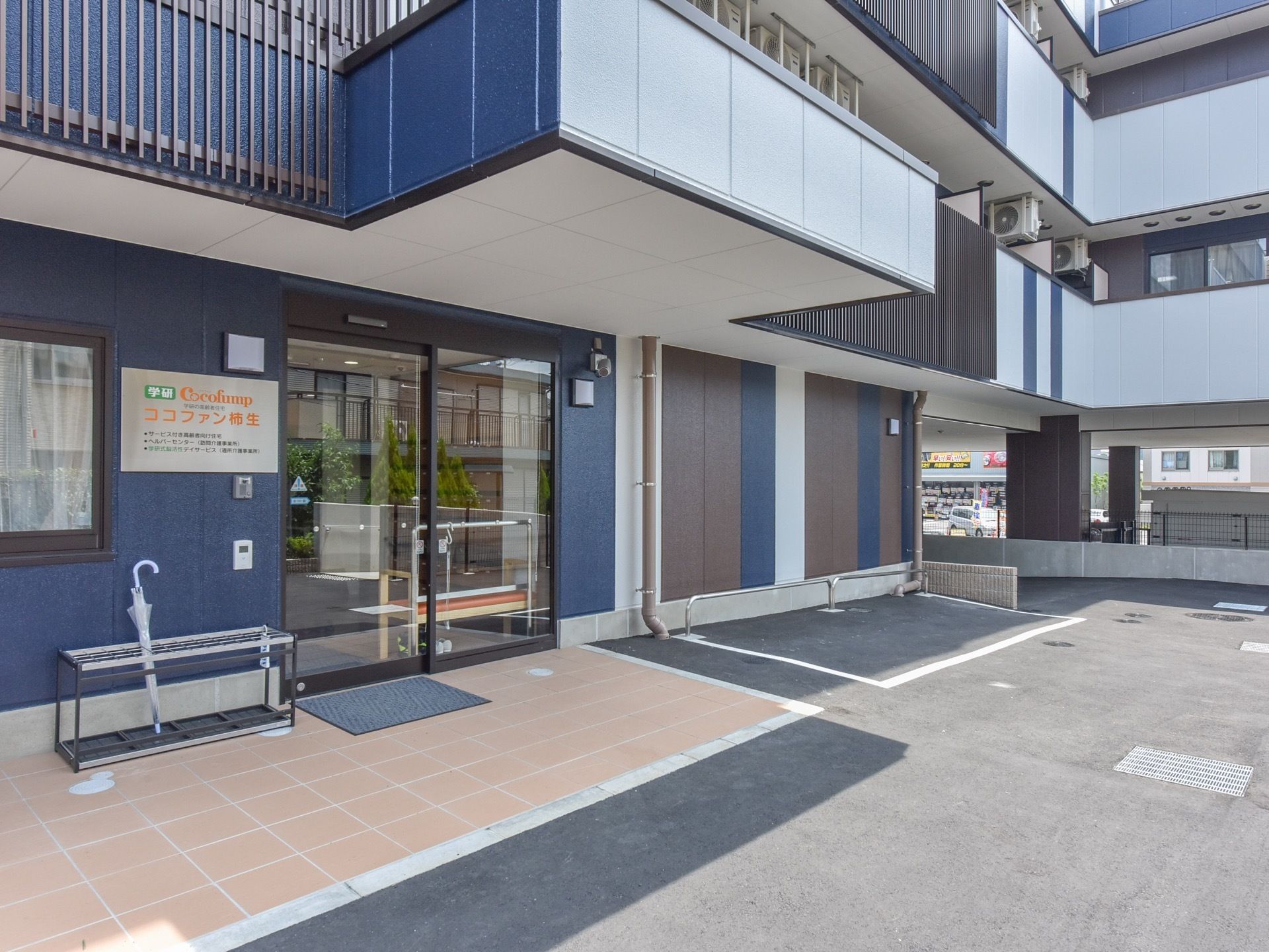 出入り口は自動ドアになっており、上にはひさしがある。側には傘立てが置かれていて、壁には施設名が書かれたプレートがついている。