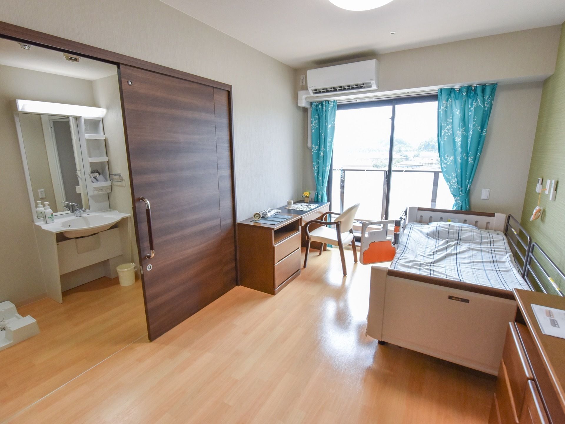 大きな窓から光が入る明るい部屋になっている。介護ベッドや机、椅子などが置かれ、ドアの向こうには洗面台が見える。